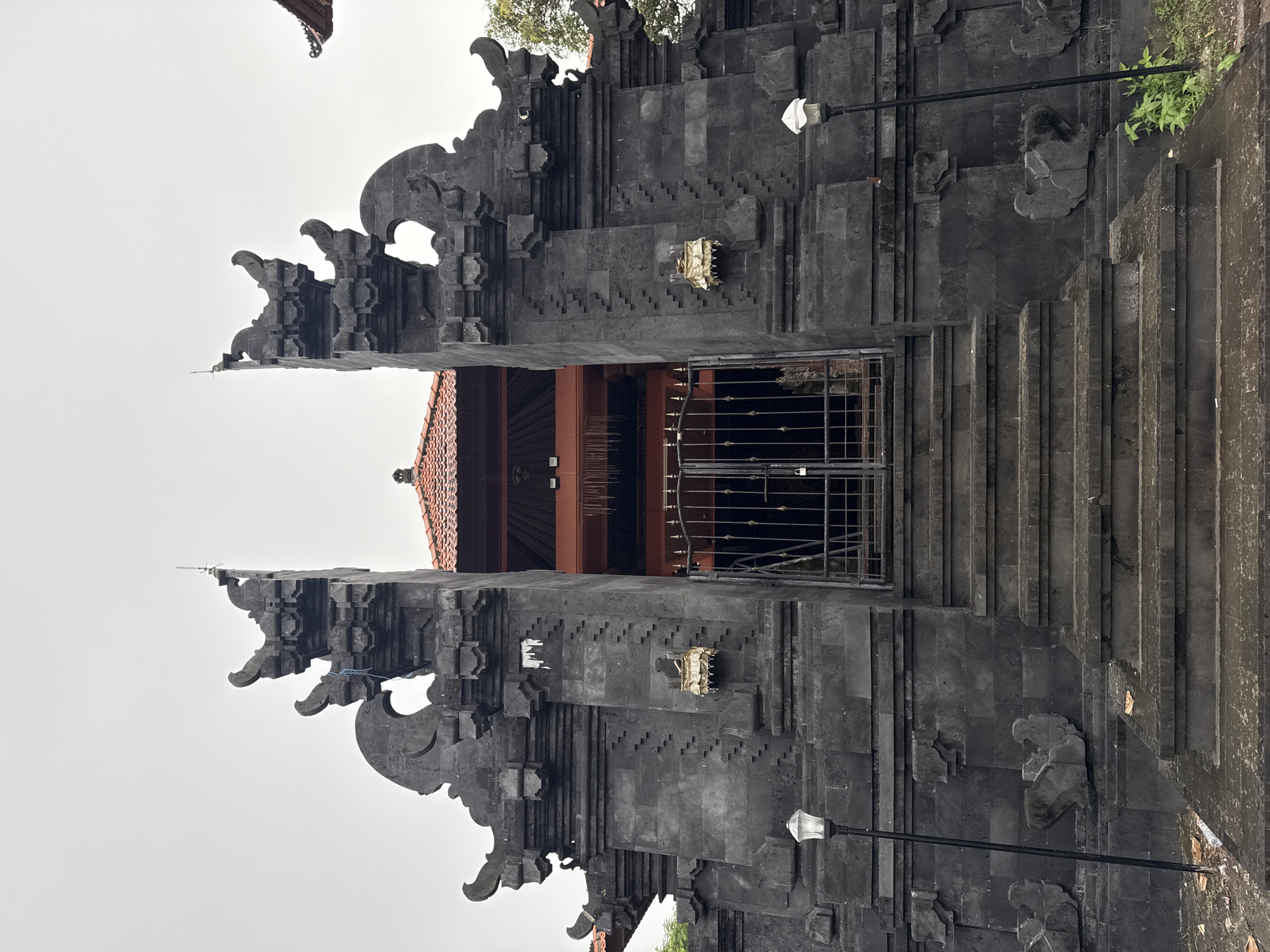 A wide, upright shot captures a grand Balinese split gate, known as a Candi Bentar, made of dark grey or black volcanic stone. The gate stands impressively with intricate carvings and traditional Balinese ornamentation on both of its tall, symmetrical halves. A broad set of stone steps leads up to the gate's entrance.
In the central opening of the split gate, a sturdy black metal gate or fence secures the passage. Behind this, a building with a prominent red-orange terracotta tiled roof and dark wooden structural elements is partially visible. Two modern-style lampposts with white, fabric-like covers over their light fixtures are positioned on either side of the lower gate structure.
Small, light-colored decorative elements or offerings are placed in niches on the stone facade. To the right of the steps, some green foliage grows on a raised section of ground. The sky is uniformly bright and overcast, suggesting a cloudy day with diffused light, likely daytime. No people are visible, and the scene depicts a static architectural view. The distinctive architectural style strongly indicates a location in Bali, Indonesia, consistent with the provided context of Kuta Selatan. There is no visible text in the image.