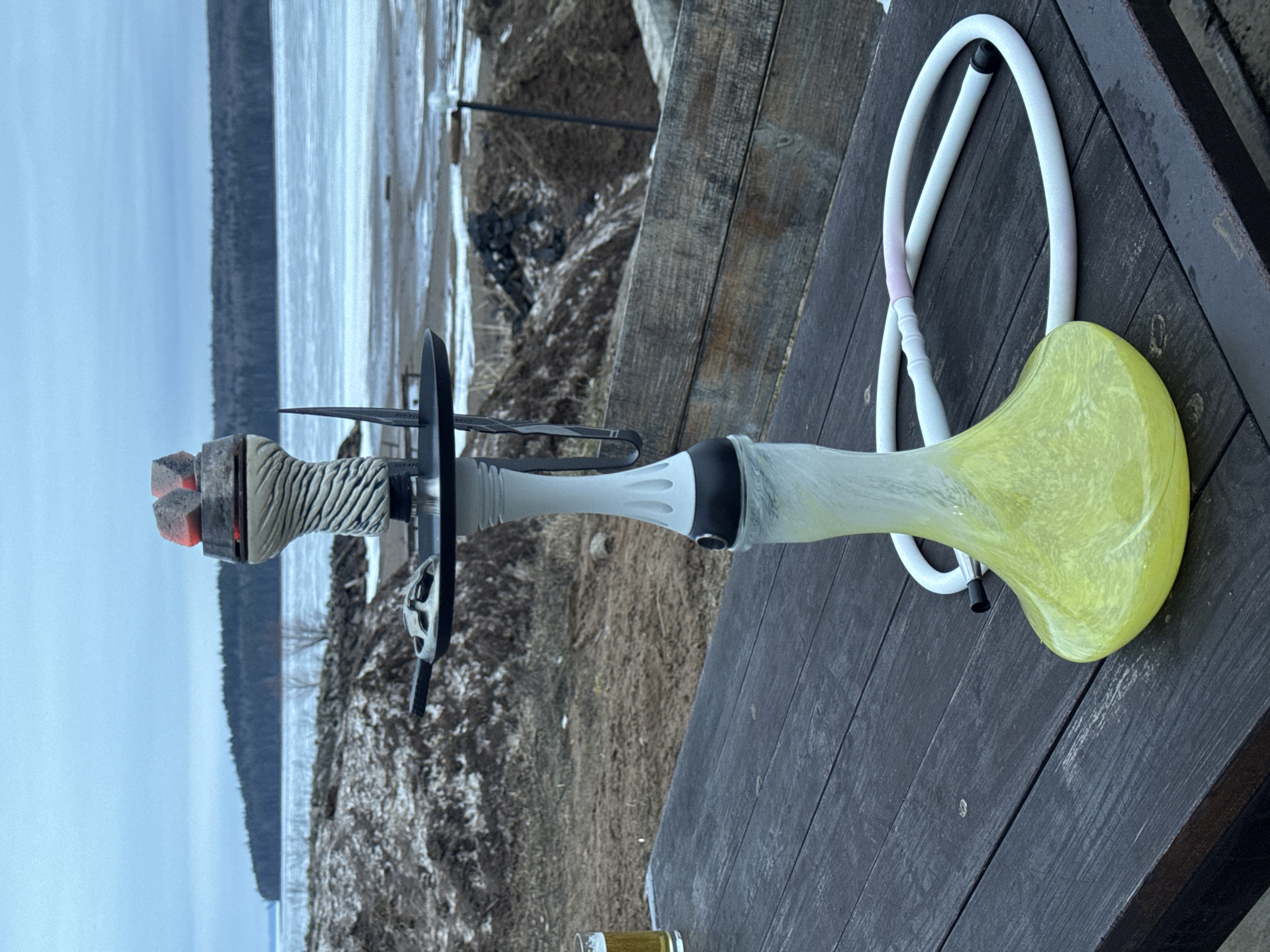 The image presents a hookah resting on a dark, weathered wooden surface. The hookah features a yellow glass base, a light-colored stem with some black accents, and a textured bowl containing what appears to be burning coals. A white hose is connected to the base.
The background shows a lakeside scene with overcast weather. Vegetation and some snow patches are visible along the shoreline.
The location is Chaikovsky, Russia.