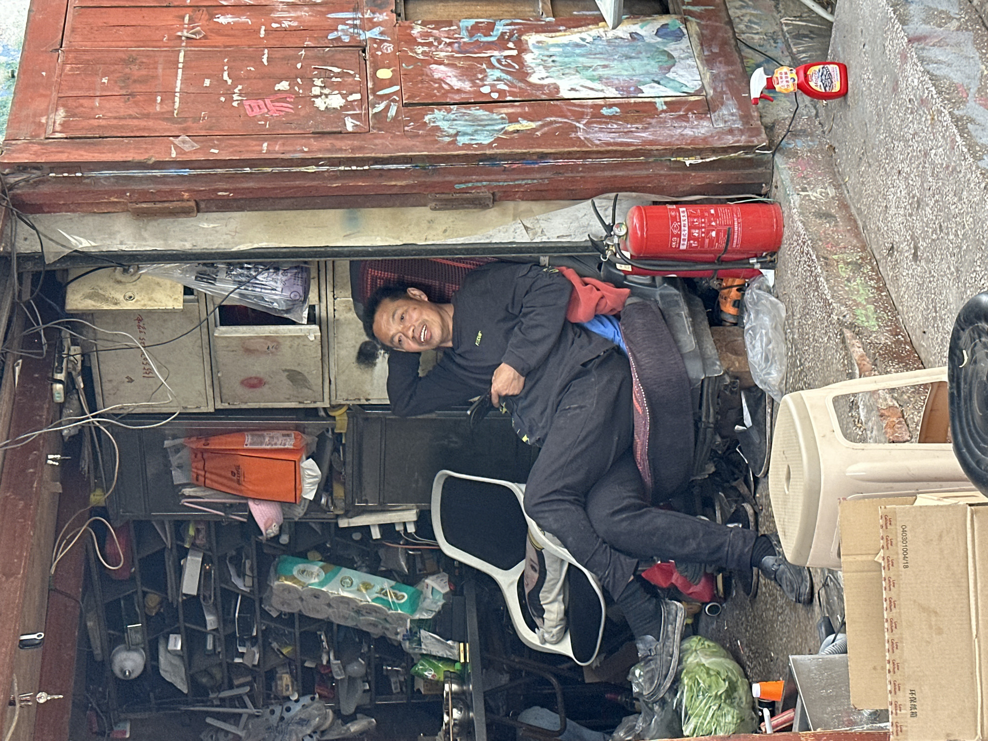 A middle-aged Asian man wearing black clothing and shoes reclines among objects crammed into a narrow space. He is smiling, looking at the camera. Above him is an old red door with peeling paint and Chinese characters. A red fire extinguisher and a bottle of cleaner sit on the ledge beside him. The cluttered space contains metal shelving, cardboard boxes, rolls of toilet paper, orange plastic bags, and other various items. Two white plastic chairs and a black office chair are also visible. Printed text is seen throughout the image, including product information and phone numbers. The city is Jiulongpo District, China.