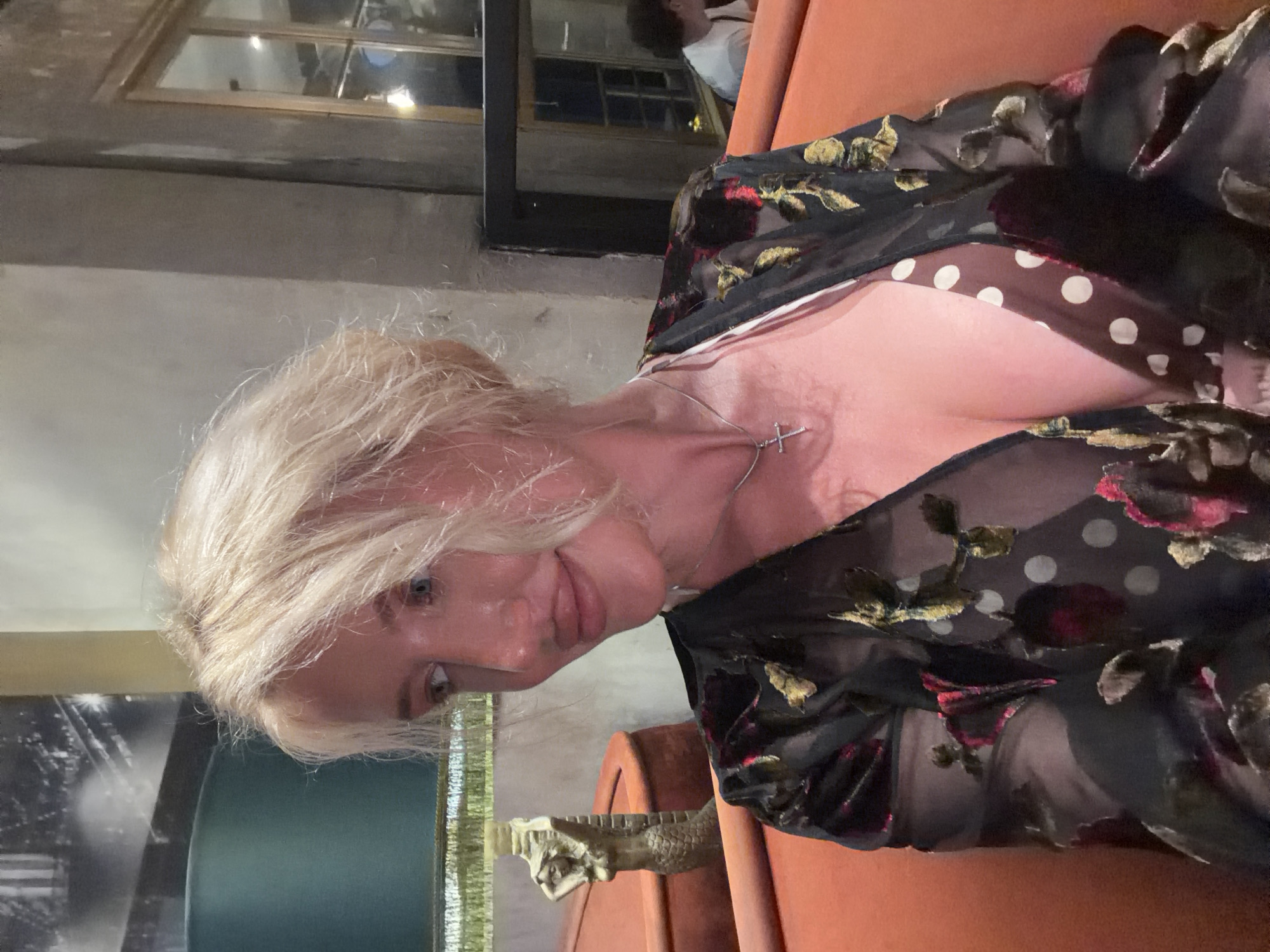 The image is a medium shot of a fair-skinned woman with short, blonde, messy hair, taken indoors. She is sitting on an orange couch, her body angled slightly away from the camera. She is wearing a dark, sheer, long-sleeved dress with a floral and polka dot pattern, layered over a light-colored camisole or top. A silver cross necklace is visible around her neck. Her face is turned towards the camera, with her lips slightly parted and her eyes looking to her left. The background appears to be a dimly lit indoor setting, possibly a restaurant or lounge, with hints of a large framed picture on the wall to the left and a window in the background to the right. The overall lighting suggests an evening or indoor ambient light setting.