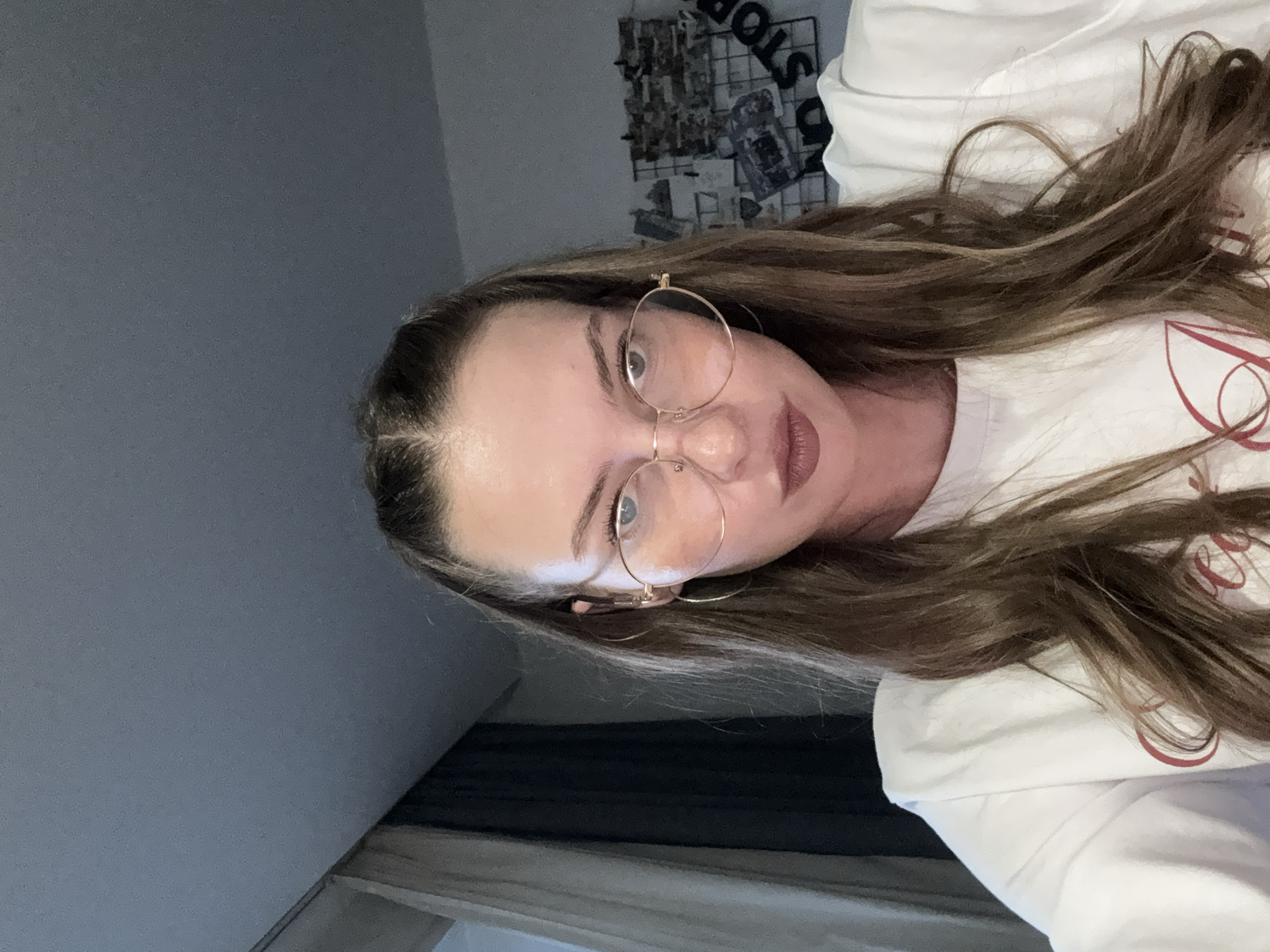 A young woman is lying down and looking at the camera. She has long, wavy brown hair, fair skin, and is wearing large, round, gold-rimmed glasses and a white t-shirt. Her makeup includes lip gloss and mascara. The background is dimly lit and appears to be a bedroom. To the left, a grey wall is visible. To the right, part of a white shirt is visible with a red design on it. Above and behind the woman, a wire grid with photos is attached to the wall. The overall mood is casual and relaxed. The image appears to have been taken indoors, likely in the evening, given the lighting. There is no discernible weather or specific location cues beyond the inferred indoor setting.