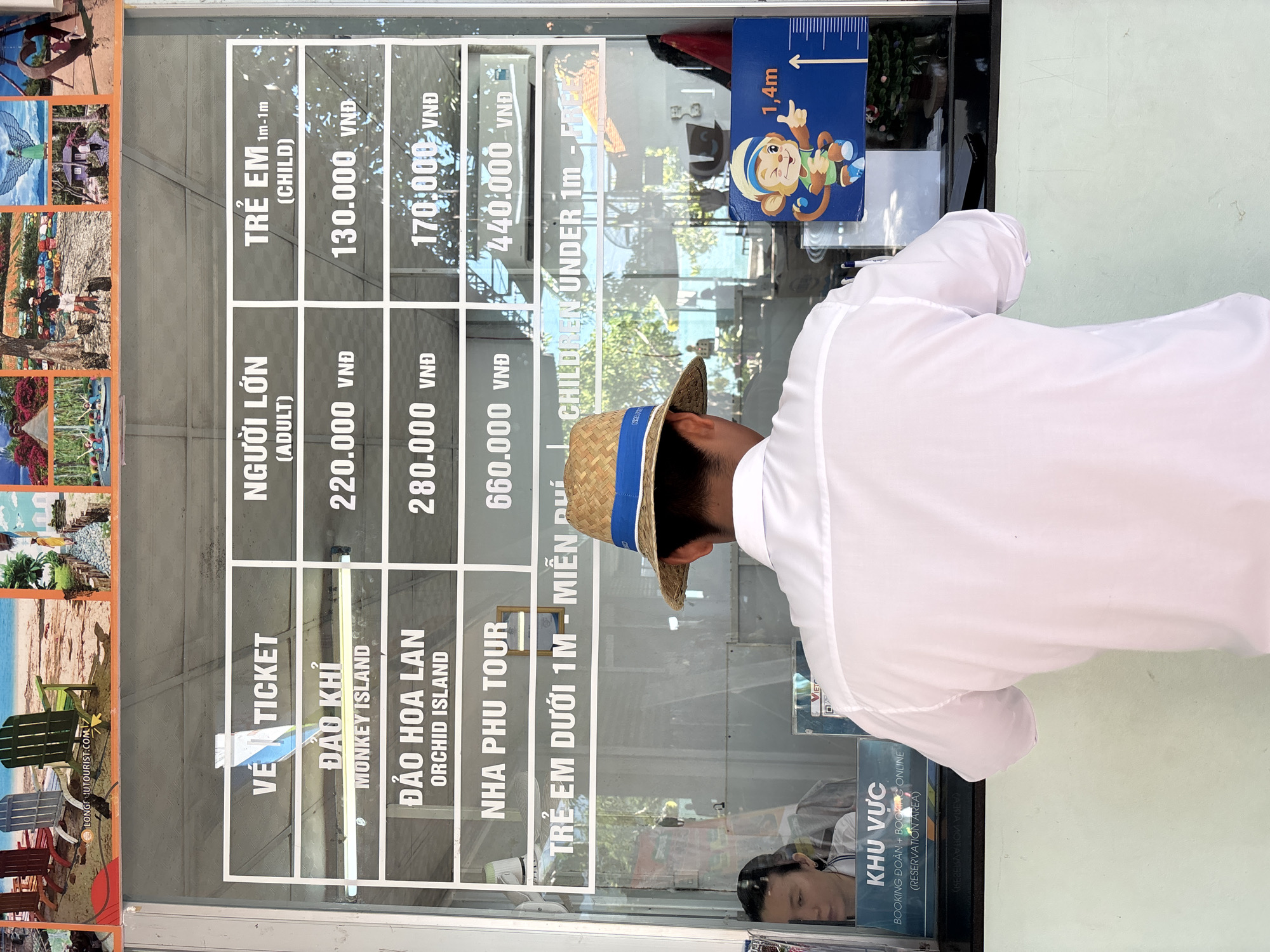 In an outdoor ticket booth, a person wearing a white shirt and a straw hat faces away from the camera, looking at a price list displayed on a glass window. The price list, visible through the glass, shows different tour packages and their corresponding prices for adults and children. The tour packages include "Dao Khi Monkey Island," "Dao Hoa Lan Orchid Island," and "Nha Phu Tour." Prices are listed in Vietnamese Dong (VND). To the right of the price list, a graphic depicting a monkey with a height measurement chart indicating "1.4m" is attached to the window. In the background, through the glass, a beach scene with lounge chairs and tropical foliage is visible. A woman is partially visible inside the ticket booth to the left. The overall environment appears to be a tourist destination, possibly a beach resort.