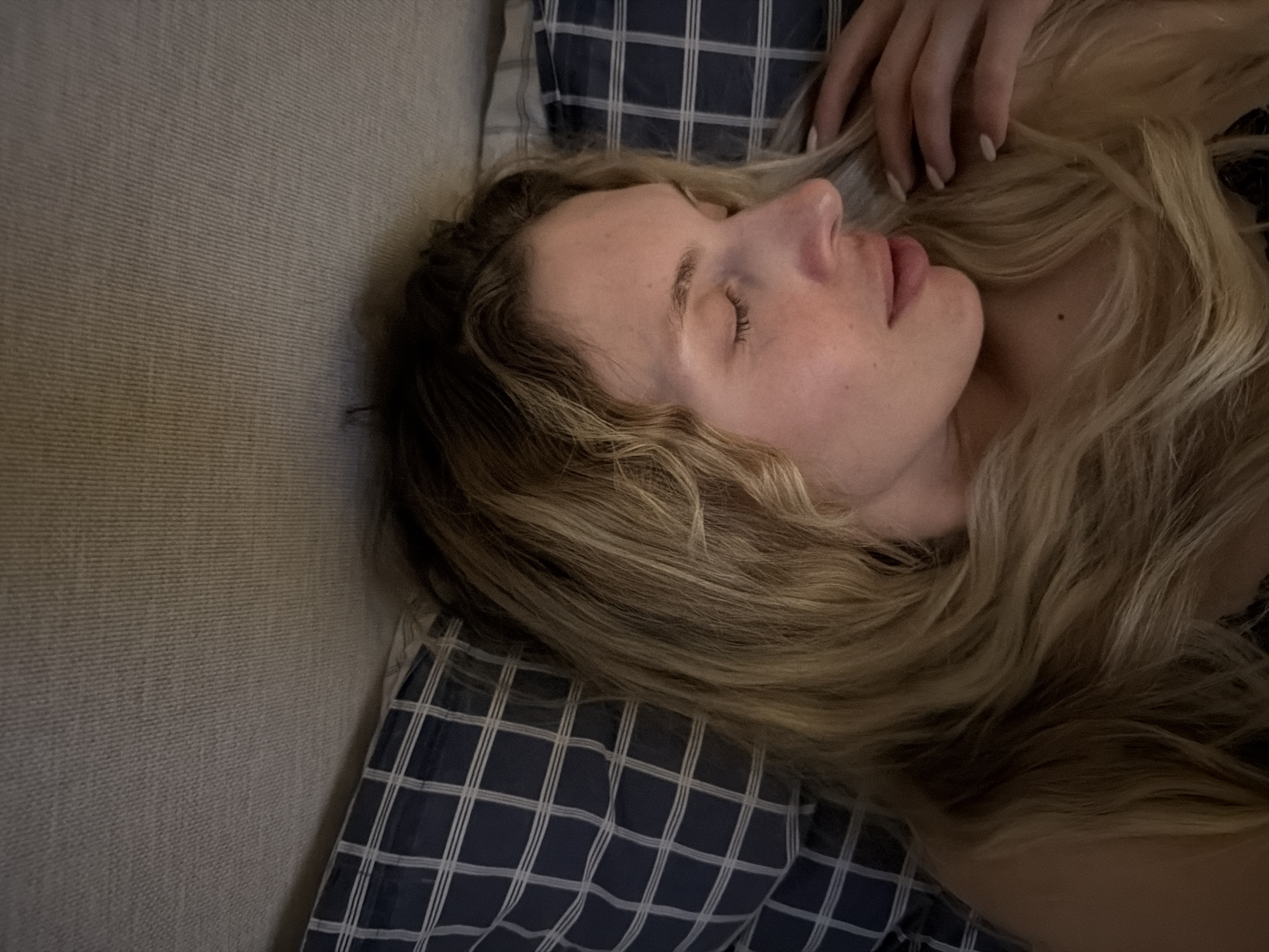 A woman is lying on her side on a gray couch with a blue and white checkered pillow. Her eyes are closed, and she has long, blond hair. One of her hands is near her head. The location is unknown and cannot be determined from the image alone. There is no indication that the location is Budva, Montenegro.