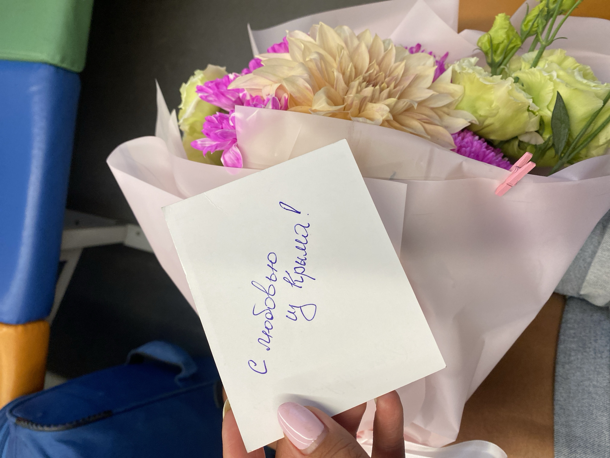 A person's hand with a light pink manicure holds a white card with blue handwritten text, placed in front of a bouquet of flowers. The flowers are predominantly light yellow and pink, with some green accents. The card appears to be tucked into the bouquet's wrapping. The background is blurred, but suggests an indoor setting with colorful seating, possibly a waiting area or public transport. The text on the card reads "С любовью из Крыма!", which translates to "With love from Crimea!". The location is identified as Vladislavovskoye rural settlement, Ukraine.