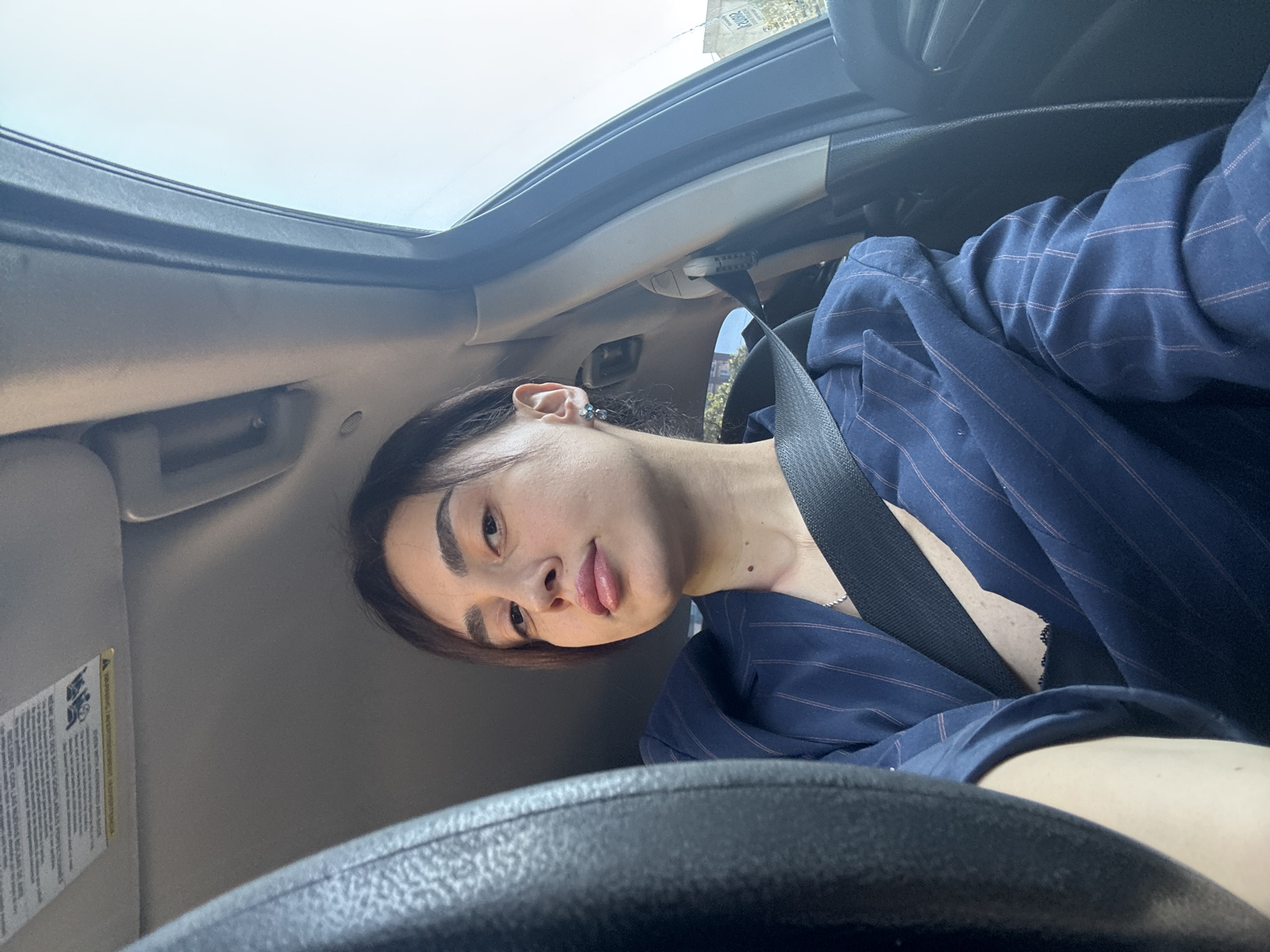A person is in a car. They are wearing a blue pinstripe blazer and a seatbelt. The car appears to have a sunroof. The location is not discernible from the image.