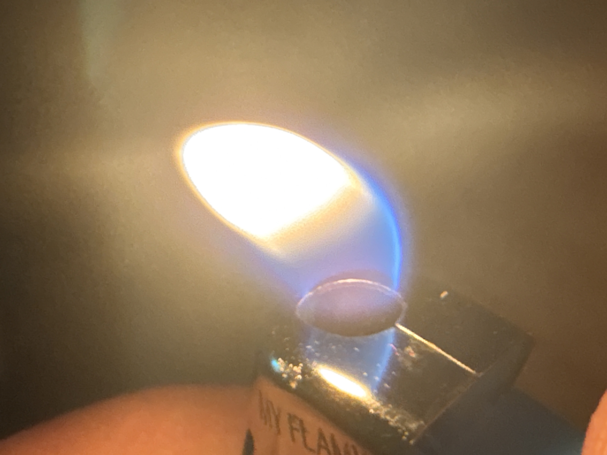 The image is a close-up, macro shot of a lighter with its flame ignited. The lighter appears to be metallic and has some text on its side, partially obscured, but "ANY FLAM" is visible. The flame is bright yellow and orange at its core, transitioning to a vibrant blue at the base, indicating it is a butane flame. There is a small, oval-shaped object, possibly a component of the lighter, visible within the flame. The background is out of focus and appears to be a muted, warm-toned surface. A portion of a person's thumb, with an orange hue, is visible on the left side of the frame, holding the lighter. The scene is indoor, with artificial lighting contributing to the illumination and reflections. There is no indication of Plovdiv, Bulgaria, or any urban environment within the image itself.