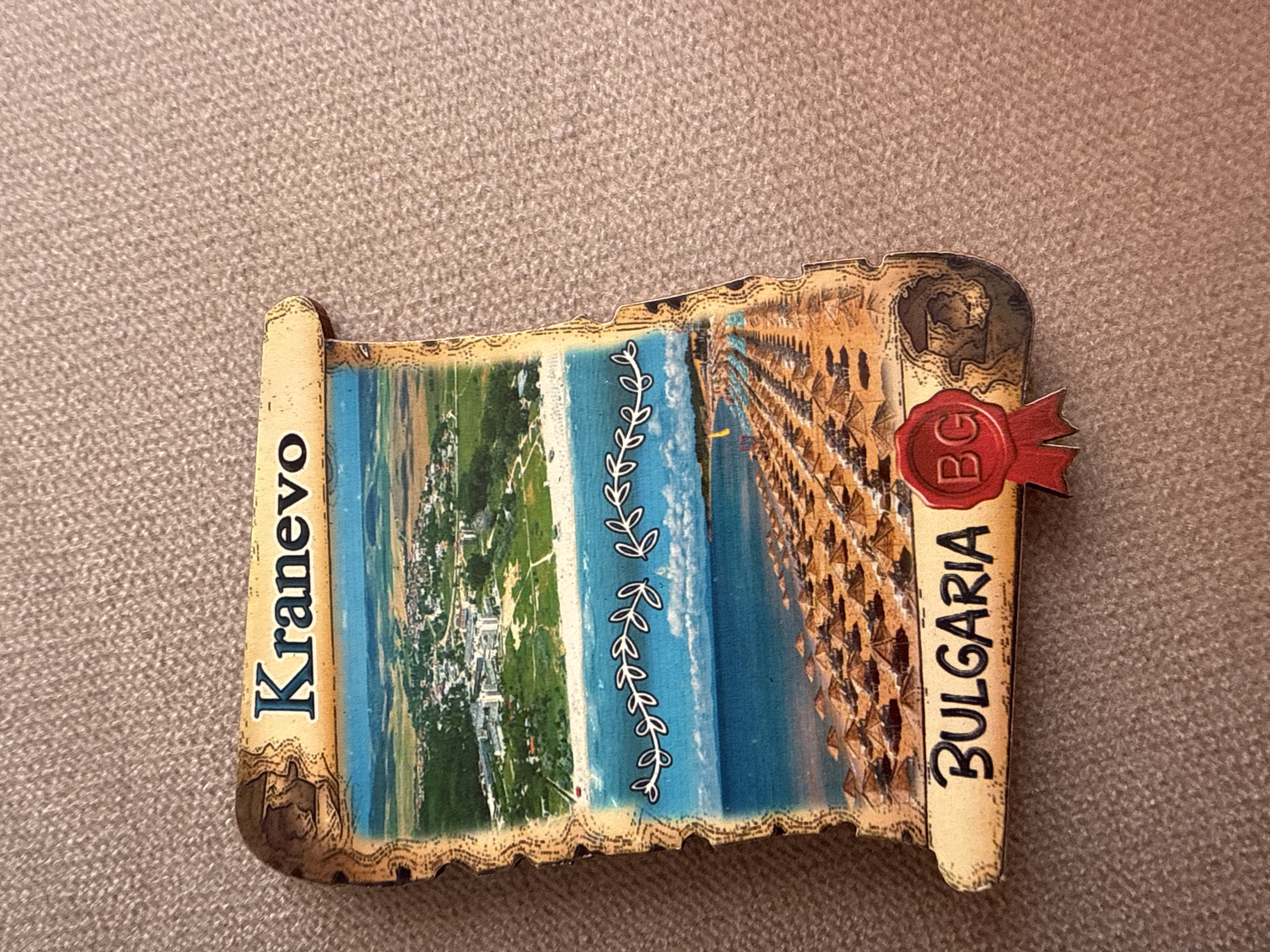 The image features a rectangular souvenir item, likely a magnet, designed in the shape of an unfurled parchment scroll. This scroll displays a vibrant, high-angle view of a coastal scene. On the left, a green landscape with numerous buildings indicates a town or resort area. This transitions into a narrow strip of white and light blue, possibly a beach or very shallow water, adorned with a stylized vertical vine pattern in white. Further to the right, a broad expanse of deep blue sea is depicted. The rightmost section of the scene shows a densely packed sandy beach lined with numerous brown parasols and sun loungers, suggesting a popular and busy tourist destination.
The "parchment" edges of the souvenir are rolled and textured, colored in a warm, antique beige. Along the left edge, the name "Kranevo" is written vertically in dark blue capital letters. On the right edge, the word "BULGARIA" is similarly oriented. A red, circular wax seal-like emblem, complete with a small ribbon, is attached to the upper right section of the scroll, displaying the white letters "BG." The item rests on a textured, light brown or beige background, possibly a carpet or fabric. The depicted scene suggests a sunny daytime setting, characteristic of a bustling beach resort.