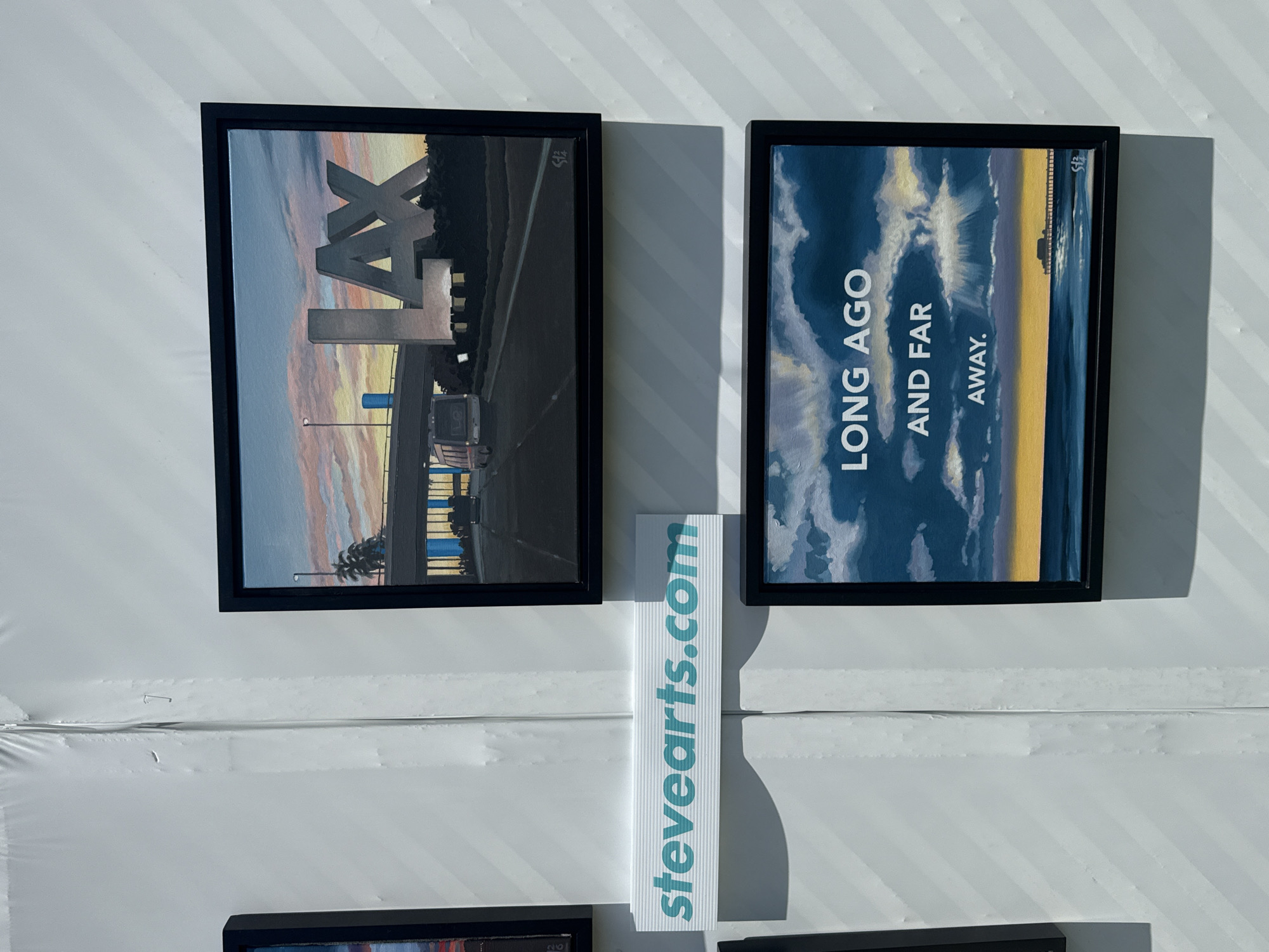 The image shows two framed paintings on a white wall with diagonal stripes. The painting on the left depicts the LAX sign with a highway and sunset in the background. The painting on the right features the text "LONG AGO AND FAR AWAY" in a vertical format, along with an image of a cloudy sky and a hint of a coastline. A white sign with the text "stevearts.com" in blue is placed between the two paintings. The location is likely an art exhibit or gallery, but there are no specifics visible that would indicate it is located in Beverly Hills, United States of America.