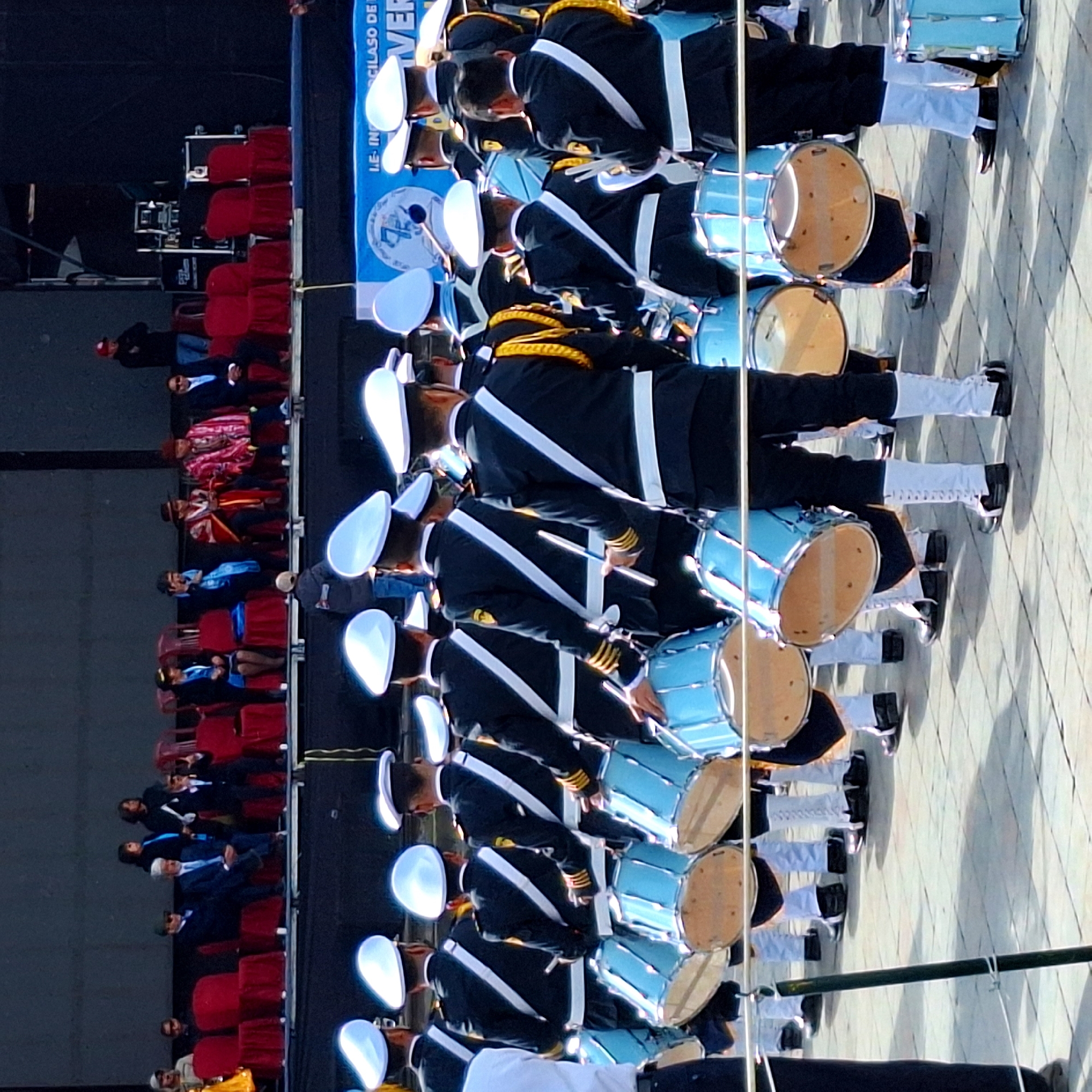 The image shows a marching band comprised of individuals in uniform, predominantly wearing dark blue jackets with white accents and white hats, carrying light blue drums. The band members are positioned on a paved surface, likely a street or plaza, with a stage in the background. Seated on the stage are spectators and officials, some wearing traditional clothing. The scene appears to be part of a public event or parade, possibly a civic or military ceremony. The backdrop includes banners and stage equipment, suggesting an organized outdoor performance. The overall atmosphere is festive or commemorative, indicated by the coordinated attire and the presence of a large gathering. The city of Cuzco, Peru, is known for its vibrant cultural events and parades, often held in its historic plazas, which could be the location depicted.