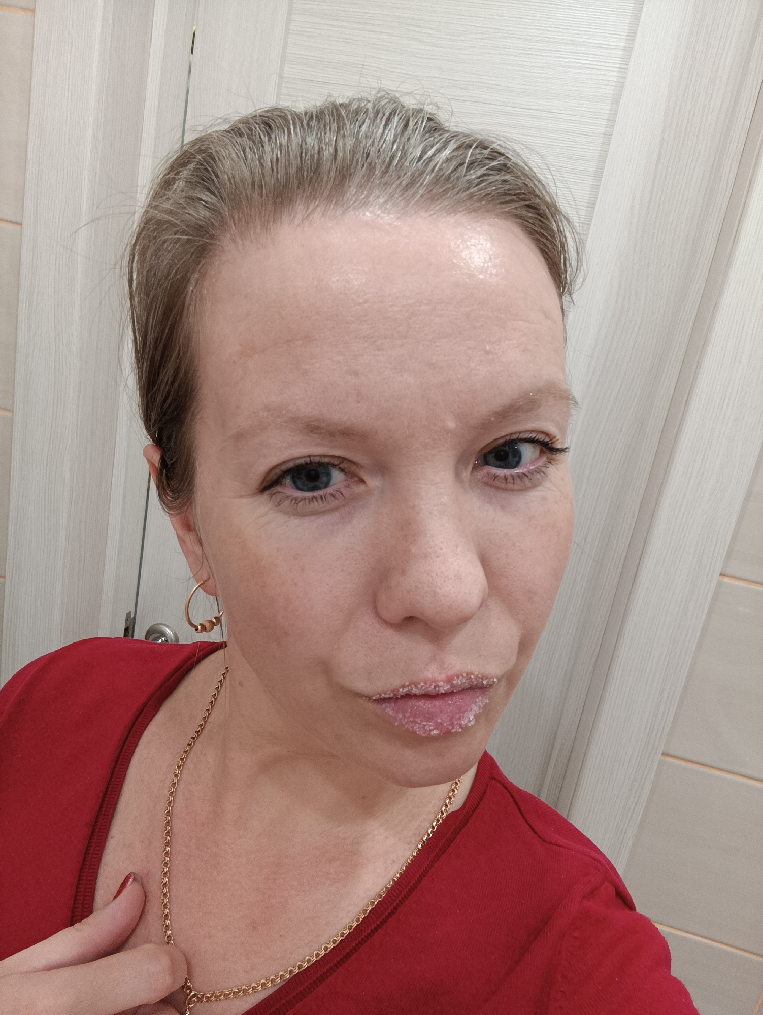 A close-up, eye-level shot depicts a fair-skinned woman with light blue eyes and short, greyish-blonde hair. She is wearing a red crew-neck shirt and gold jewelry, including hoop earrings and a delicate chain necklace. Her lips are covered in a white, crystalline substance that resembles sugar or salt. The background features a white door with vertical panels. The location is identified as the city of Chaikovsky, Russia. The image appears to be a selfie taken indoors.