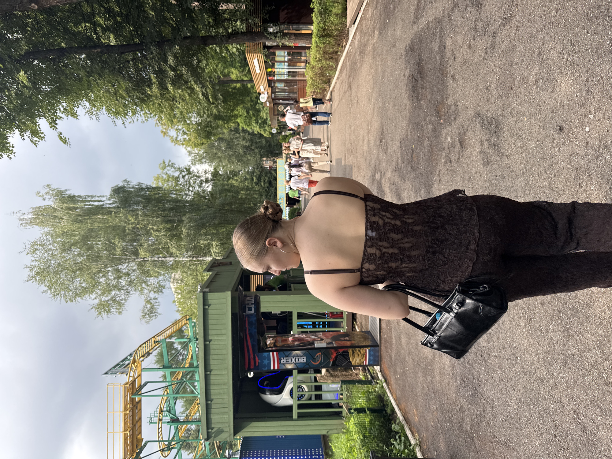 The image depicts a woman walking away from the camera on a paved path in what appears to be an amusement park. She is wearing a dark brown lace top and matching pants, carrying a black handbag. To her left, there is a green structure with a roller coaster visible in the background. Various stalls and trees line the path, with other people walking in the distance. The sky is overcast with clouds. This scene is located at the Amusement Park "Park Pobedy" (Victory Park) in the city of Perm, Russia.