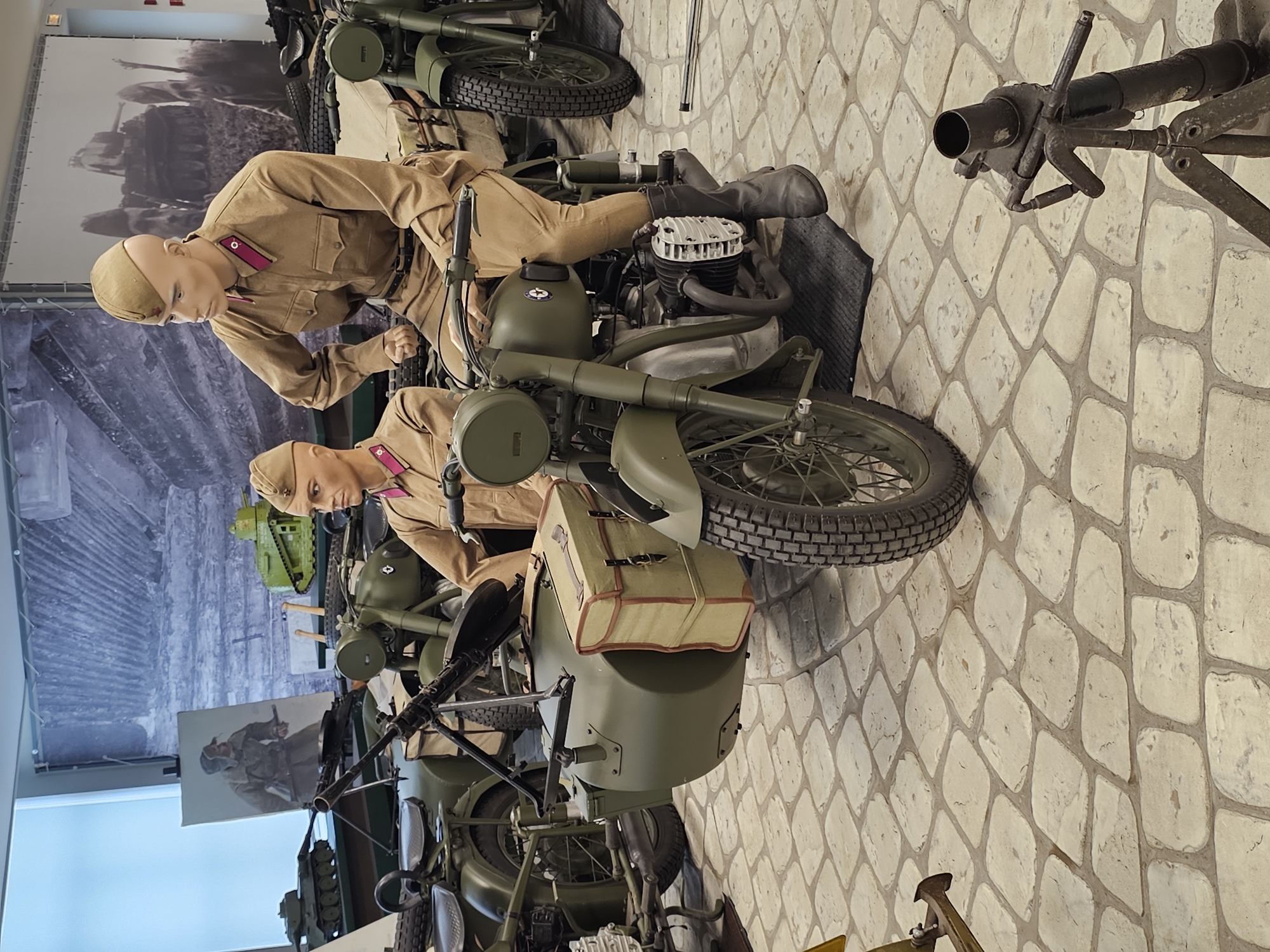 The image depicts an exhibit in Архангельское, Russia, featuring vintage military motorcycles with sidecars. Two mannequins dressed in period military uniforms are positioned on the motorcycles, simulating riders. The motorcycles are olive green and appear to be from the World War II era or shortly after. In the background is a large mural depicting soldiers in battle. The floor is a light-colored stone pattern. A mortar is situated on the right side of the image.