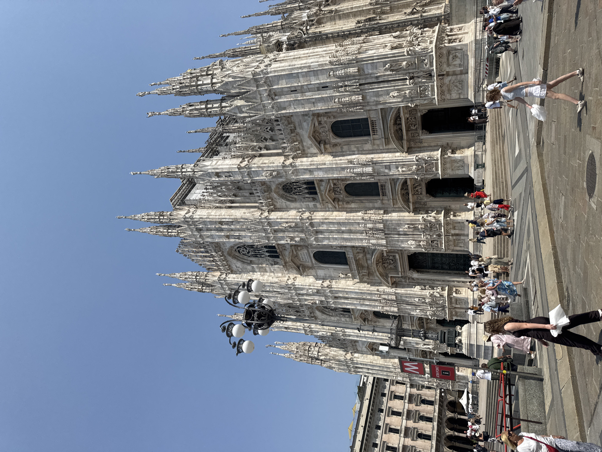 The image shows a wide-angle, eye-level outdoor shot of the Milan Cathedral, also known as Duomo di Milano, in Milan, Italy. The cathedral, a massive Gothic structure made of white marble, dominates the background, with its intricate spires, statues, and ornate facade. The sky above is a clear, bright blue.
In the foreground and mid-ground, a public square is bustling with activity. Numerous people, dressed in casual summer attire, are walking, standing, and interacting. Some are tourists taking in the sights, while others appear to be locals going about their day. There are also elements of urban infrastructure visible, including street lamps, a subway entrance sign (indicated by a red 'M' logo), and some red benches in the left background. The ground is paved with cobblestones and some crosswalks. The overall impression is of a lively tourist destination on a sunny day.