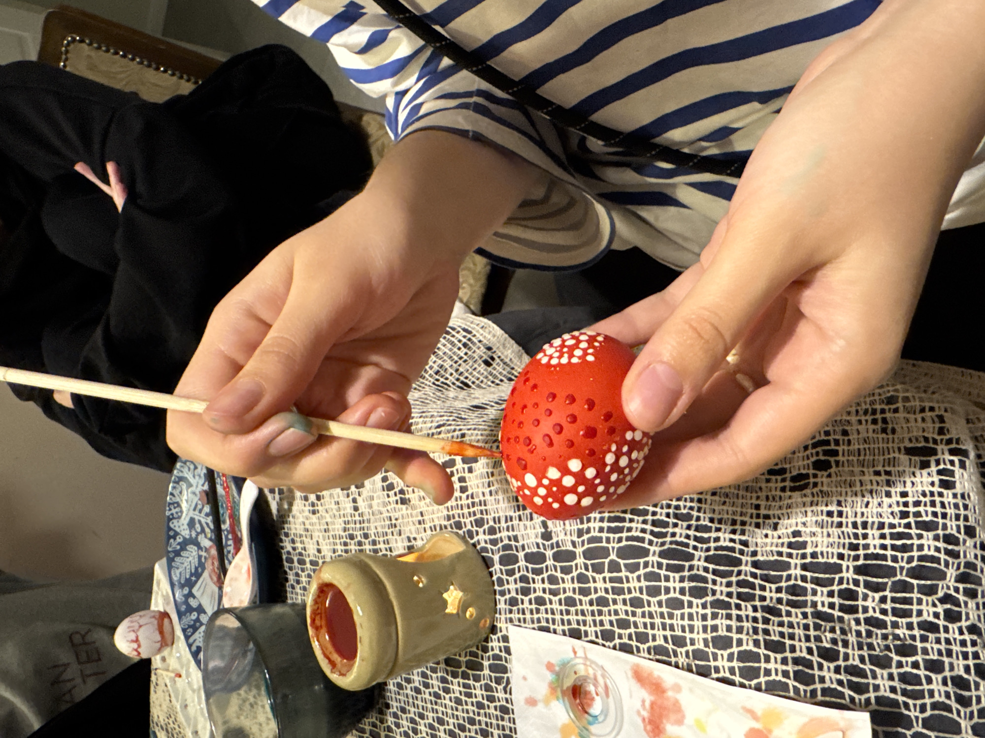 A person in a blue and white striped shirt is shown painting a red egg with white polka dots. The person is holding a thin paintbrush in their left hand, which is dipped in a small container of red paint. The red egg is being held in the person's right hand. The egg has several white polka dots already painted on it. The scene is set on a tablecloth with a crocheted or lace pattern. The exact location in Moscow, Russia is not discernible from the provided image.