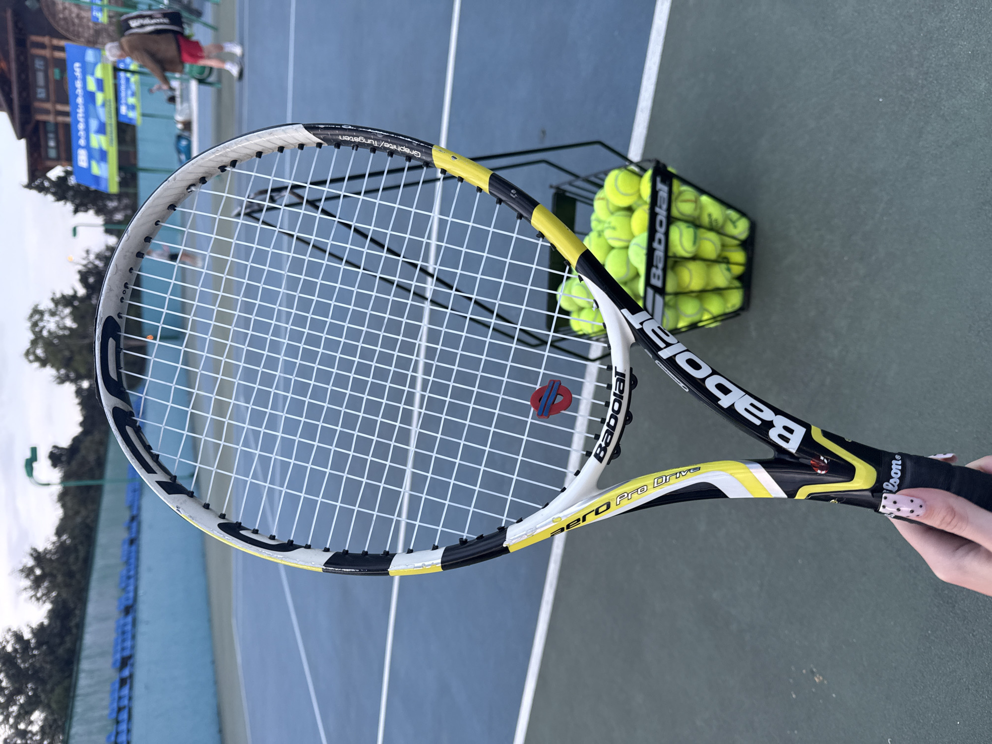 A tennis racket is held in front of a tennis court. The racket is a Babolat Aero Pro Drive, and a red and blue vibration dampener is visible on the strings. A basket full of tennis balls sits to the side of the racket. In the background, a tennis court is visible with blue and green surfaces. In the far background, signage is visible with the letters "Upse..." visible, and a person in athletic wear is standing on the court. The setting appears to be in Batumi, Georgia.