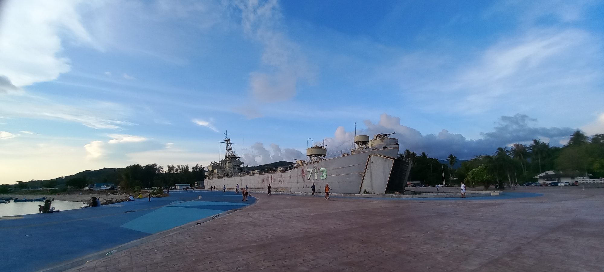 Captured in Thong Sala, Thailand, the image presents a static, gray naval vessel marked with the number "713", situated on land. The vessel, possibly a preserved or retired ship, is the focal point, surrounded by a paved area where several individuals are visible, some walking, suggesting a public space. To the left, a blue painted path borders a harbor, and in the background, lush tropical vegetation and a building are visible. The sky, a mix of blue and cloud formations, indicates an overcast or partially cloudy weather condition.