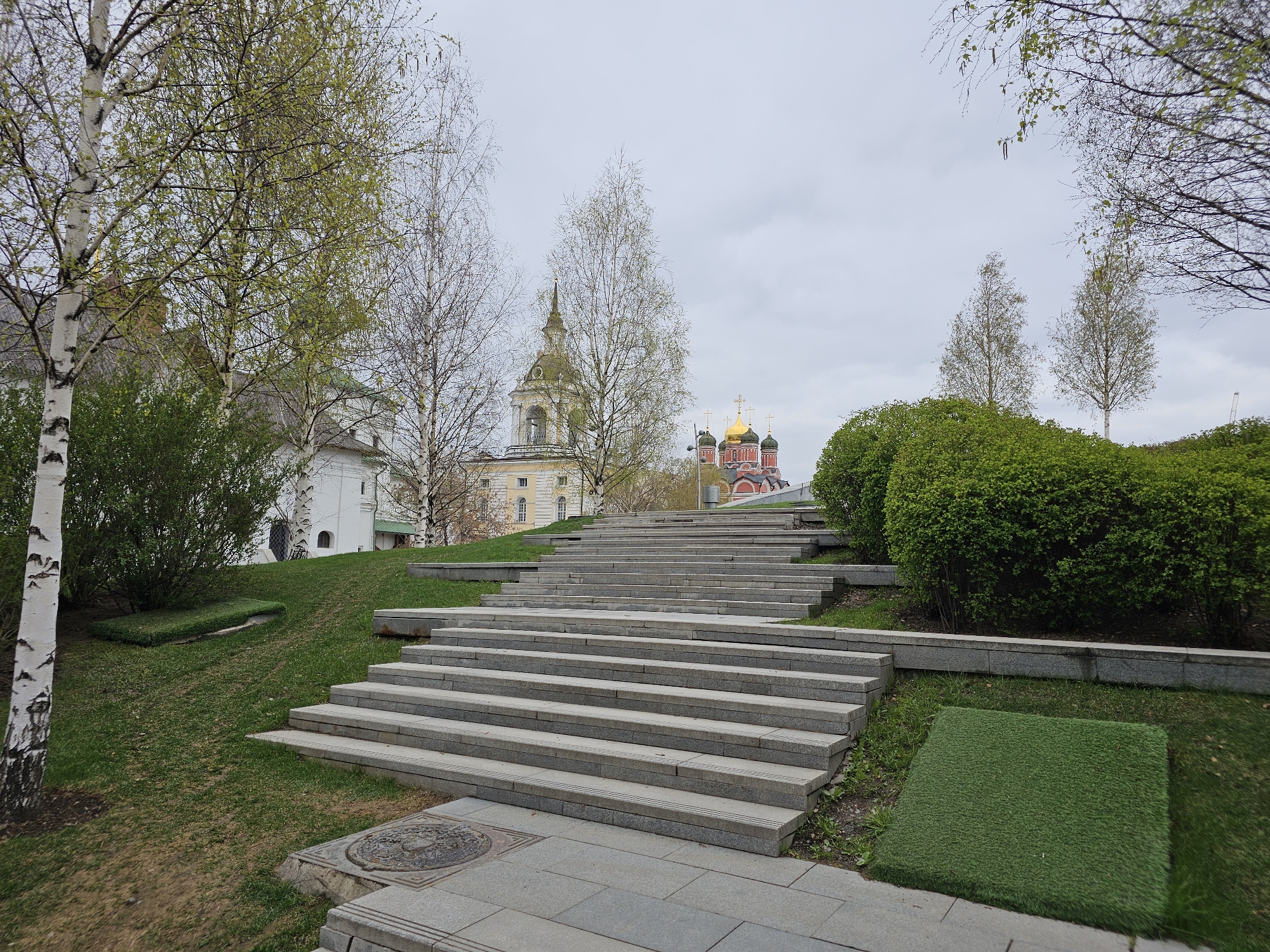 The image depicts a set of wide, stone steps leading up a grassy hill in Zaryadye Park, Moscow, Russia. The steps are bordered by green bushes and patches of artificial turf. In the background, there is an architectural view of the city, including a building with a bell tower and the domes of a Russian Orthodox church. The sky is overcast. The trees have budding leaves, suggesting it is spring. There are no people visible in the image. A tree with white bark is partially visible on the left. A circular manhole cover is visible in the foreground.