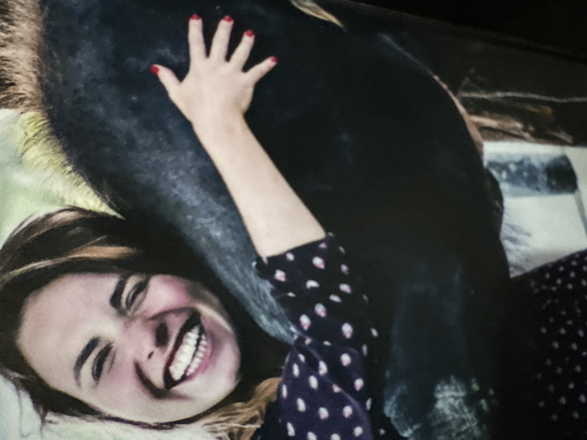 This media file is a photograph of a woman and a horse. The woman is lying down, with her head tilted back and a wide smile on her face. Her eyes are closed, and her mouth is open in a laugh. She is wearing a dark shirt with a white polka dot pattern. Her right arm is wrapped around the horse's body, and her left hand is resting on its flank. The horse is large and dark in color, with a thick mane. The background is blurred but appears to be outdoors, possibly in a natural setting. The lighting suggests it is daytime. The overall mood of the photograph is joyful and affectionate, capturing a moment of connection between the woman and the horse. The woman's red nail polish is a notable detail. The image is cropped, with the top of the horse and the full extent of the woman's body not visible. There is no visible text in the image.