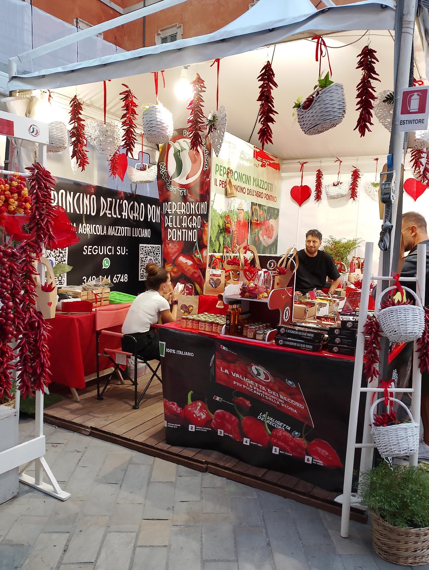 The image depicts an outdoor stall selling chili peppers, likely at a local market or festival in Rieti, Italy. The stall is decorated with numerous strings of dried red chili peppers, white woven heart ornaments, and small white woven baskets. A black banner displays text in Italian, including "DONCINO DELL'AGRO PONTINO" and "PEPERONCINI MAZZOTTI".
Two individuals are visible. A woman with her hair tied back in a bun, wearing a white t-shirt and dark skirt, is seated at a small table, looking at her phone. She is positioned on the left side of the stall. A man, wearing a black t-shirt and dark pants, stands behind a display table on the right side of the stall. He is interacting with an unseen customer or colleague.
The stall features various products, including jars of what appear to be chili-infused products and packaged goods. A white ladder-like structure is present on the right side, also decorated with chili peppers and a basket. The ground is paved with rectangular stones, and a wooden platform is visible in front of the stall. The overall impression is a vibrant and festive presentation of agricultural products.