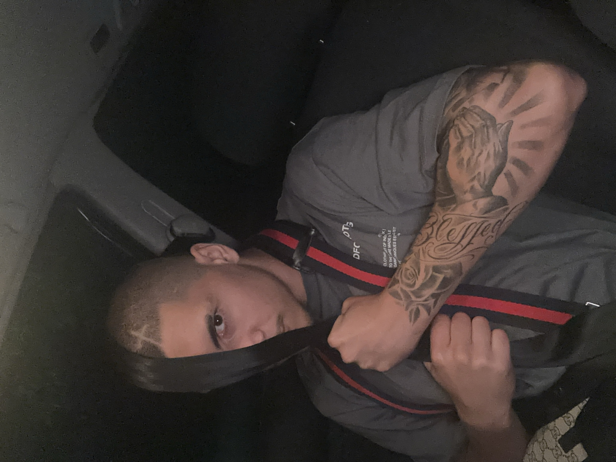 A man is lying in what appears to be the interior of a car. He is wearing a grey t-shirt and a black and red strap that crosses his body. The man has short hair with a design shaved on the left side of his head. He has a tattoo covering his left arm with images of praying hands, a rose, and the word "Blessed." He is partially obscured by a seatbelt. The background is dark, showing the car's interior. The image was taken indoors. The location is not apparent from the image and is not discernible as being in Plovdiv, Bulgaria.