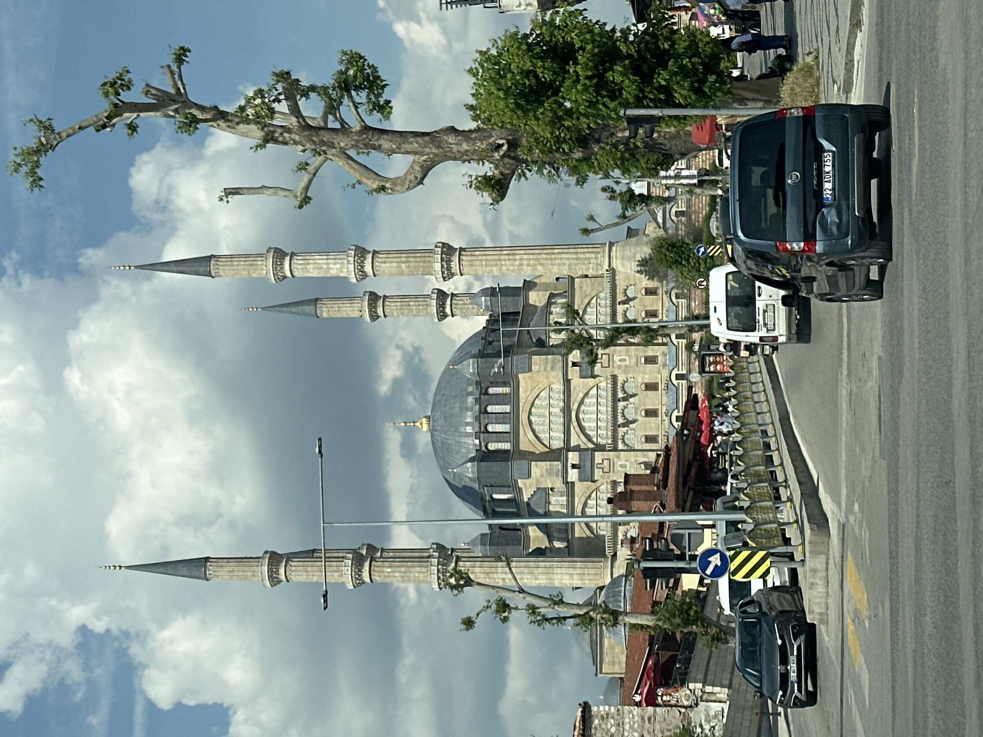 This detailed street-level image captures the Selimiye Mosque in Edirne Merkez, Turkey, under a partly cloudy sky. The central focus is the grandeur of the Selimiye Mosque, identifiable by its large central dome, surrounding smaller domes, and four tall, slender minarets, each topped with a dark, pointed spire and a golden crescent finial. The mosque's facade is constructed of light-colored stone, featuring multiple arched windows and intricate architectural details.
In the foreground, a paved two-lane road stretches away from the viewer. On the left lane, a dark grey Honda Civic, license plate "22 AET 687," proceeds forward. On the right lane, a dark teal Fiat Fiorino, license plate "22 ADK 755," also moves away; its rear bumper on the passenger side shows a visible dent. A white van with license plate "22 AAD 208" is partially visible behind the Fiat.
The road is bordered on the right by a sidewalk separated by a low, ornate golden-colored metal barrier. Various street fixtures are present, including a blue and white "keep right" arrow sign, a yellow and black striped hazard sign, and light poles with LED luminaires. A large, gnarled tree with sparse green leaves stands on the right side of the road, partially obscuring the mosque's minarets. Further along the sidewalk, several pedestrians are visible, one wearing a light blue top and dark pants.
To the left and right of the main road in the midground, other buildings with reddish-brown tiled roofs and some red awnings suggest commercial establishments or residential structures, leading up to the mosque complex. A stone wall structure is visible on the far left. The overall scene depicts a busy urban environment centered around a significant historical and religious landmark.