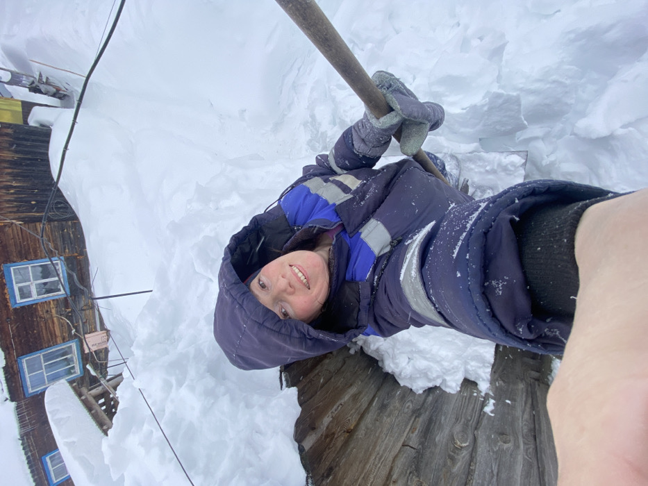 An overhead selfie captures a smiling female individual standing in a deep, snow-lined trench in Узяр, Russia. She is dressed in a dark blue winter jacket with blue and grey reflective stripes, a raised hood, and grey gloves, holding a wooden-handled shovel with her right hand. The trench walls, composed of significant snow accumulation, rise above her head. To her left, a weathered, dark wooden structure, possibly a well casing or wall, is partially visible. In the background, a rustic, two-story wooden building with light blue-framed windows is heavily covered in snow, with drifts reaching the level of its lower window sills. Several black utility wires are strung across the deep snow and near the building. The entire scene is blanketed by heavy snow, indicating a severe winter environment.