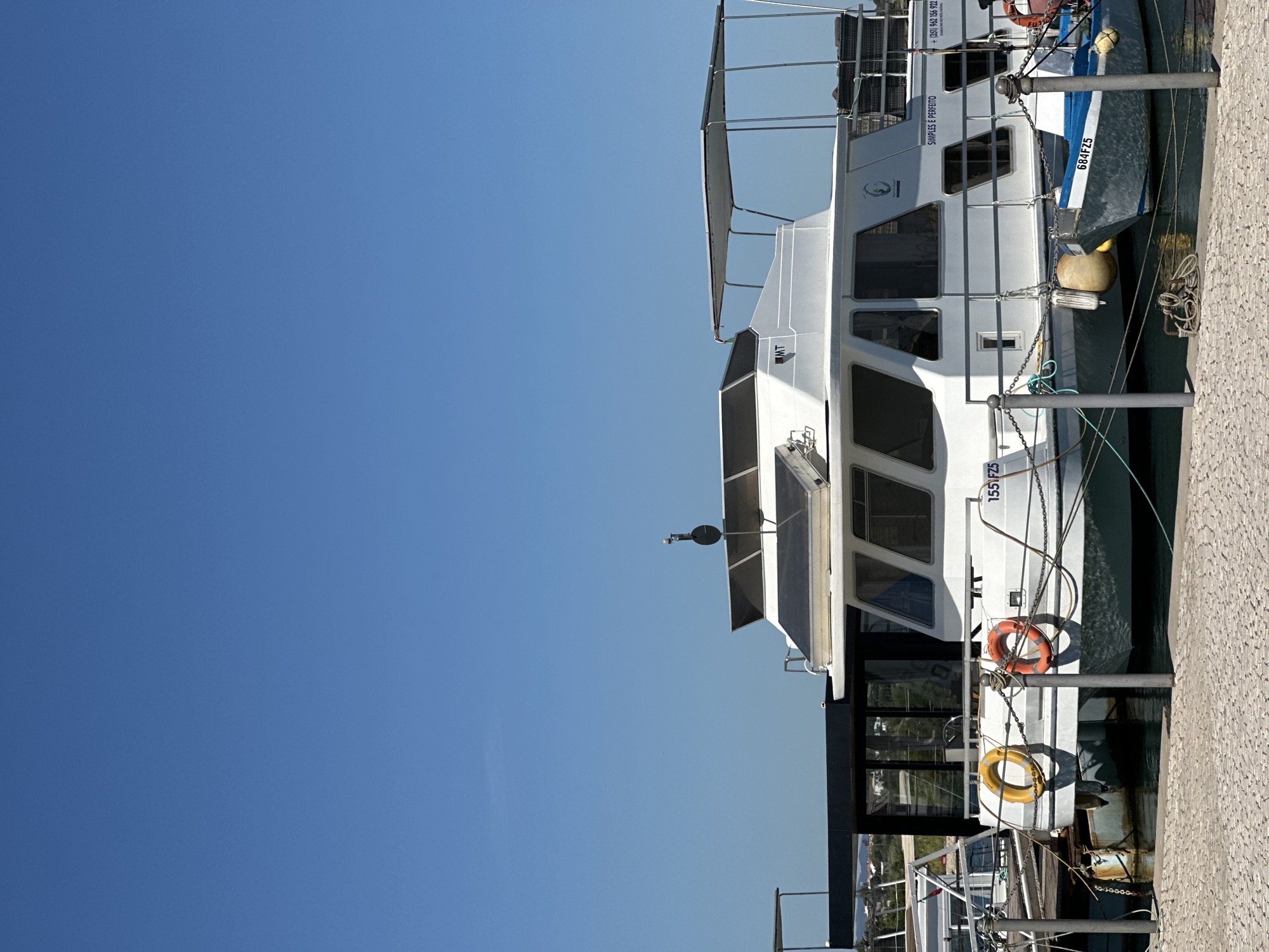 The image captures a vessel docked in what appears to be the city of Olhão, Portugal. The sky is a clear, bright blue, contrasting with the white and gray structure of the boat. Visible on the vessel are identification markings "155 FZ5" and "684FZ5," along with the text "SIMPLES E PERFEITO" and a Portuguese phone number. The boat is moored alongside a concrete walkway or dock, secured by chains.