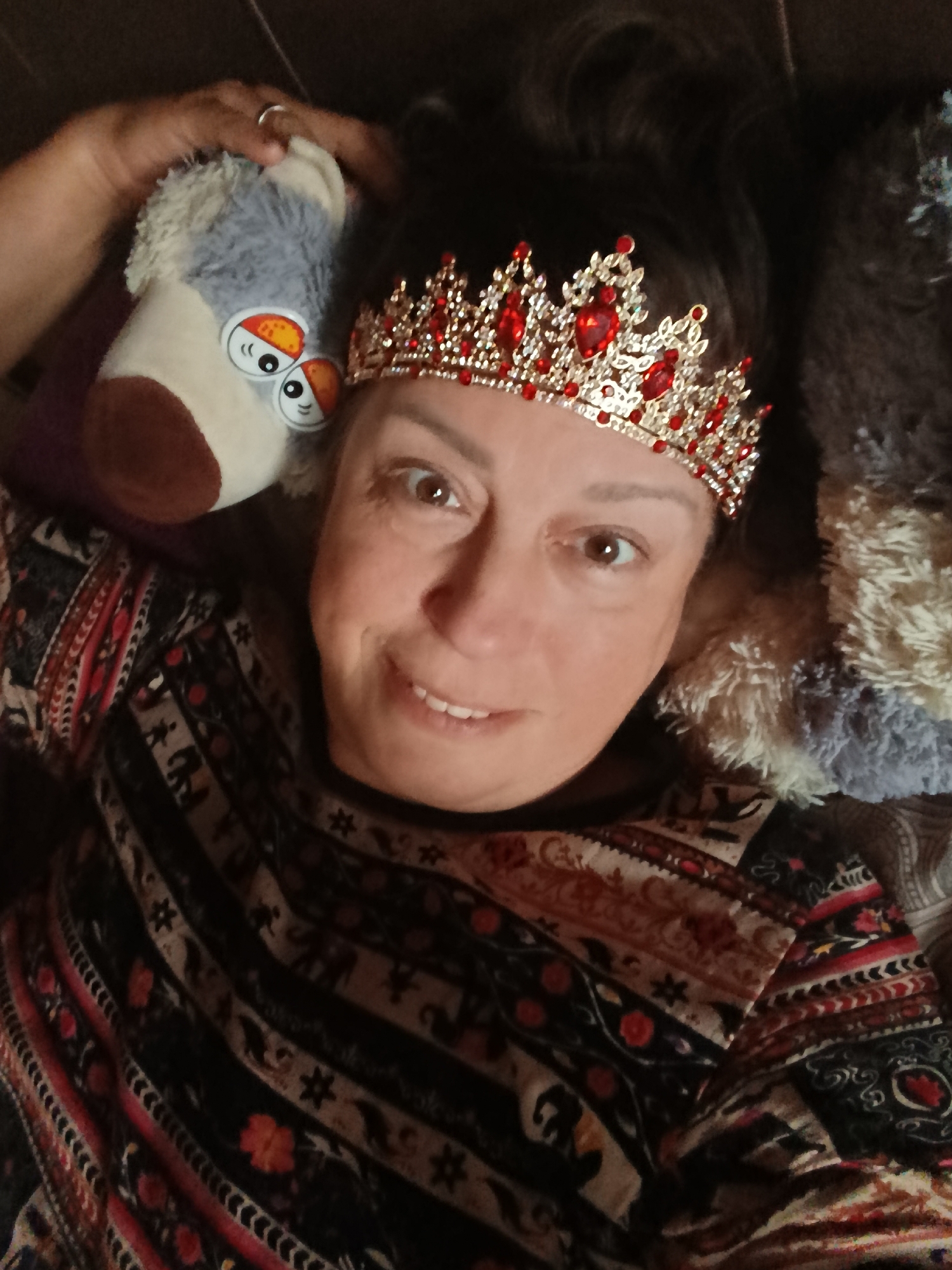 A woman with brown hair and brown eyes is wearing a red and gold crown. She is lying down on her back and smiling at the camera. She is also holding a stuffed animal in her left hand. The stuffed animal is gray and white with orange and black eyes. The woman is wearing a patterned shirt with red, black, and white designs. The background is dark and blurry. The scene is indoors, likely in a bedroom or living room. The lighting is dim.