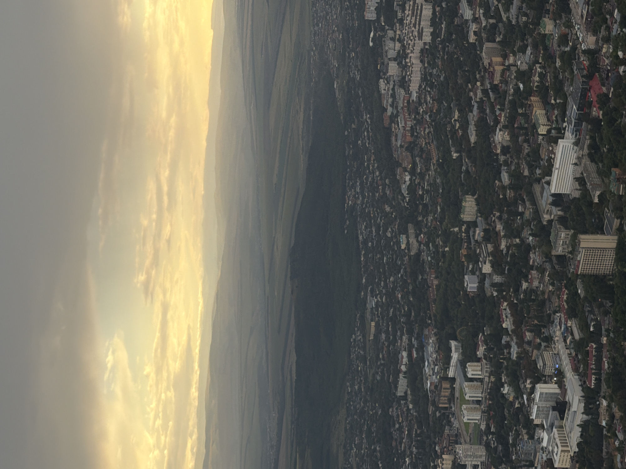 The image captures an aerial view of Goryachevodsky, Russia, during what appears to be either sunrise or sunset. The sky is filled with warm, yellowish light filtering through a layer of clouds, creating a soft, diffused illumination over the landscape. In the foreground, a dense urban area is visible, characterized by a multitude of buildings of varying sizes and architectural styles, interspersed with trees and green spaces. Streets and roads weave through the city, connecting different neighborhoods.
Further into the distance, the urban sprawl gives way to a large expanse of dark green forest, suggesting a significant wooded area bordering the city. Beyond the forest, the terrain transitions into rolling hills and mountains, softly outlined against the hazy, light-filled sky. The overall impression is one of a city nestled within a natural landscape, with the sky providing a dramatic and atmospheric backdrop. No people, specific subjects, or actions are clearly discernible in this high-altitude, broad-view shot.