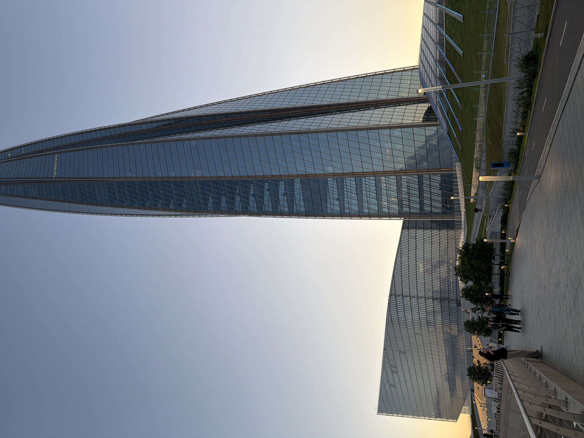 In the city of Saint Petersburg, Russia, an exterior, low-angle, full shot captures the Lachta Center skyscraper and its surrounding park area at dusk. The skyscraper, a tall, glass-covered structure with a pointed apex, dominates the right side of the frame, its multifaceted surface reflecting the pale blue of the sky. To the left of the skyscraper, a lower, angular building with a glass facade mirrors the skyscraper's modern aesthetic. A paved walkway leads into the foreground, with several individuals walking and standing on it. To the right of the walkway, a grassy incline is illuminated by blue lights, suggesting a decorative or functional lighting system. The sky transitions from a clear blue at the top to a soft yellow-orange near the horizon, indicating the sun has recently set. Trees are scattered across the landscape, adding a touch of nature to the urban setting. A railing runs along the left edge of the walkway, separating it from a lower level.