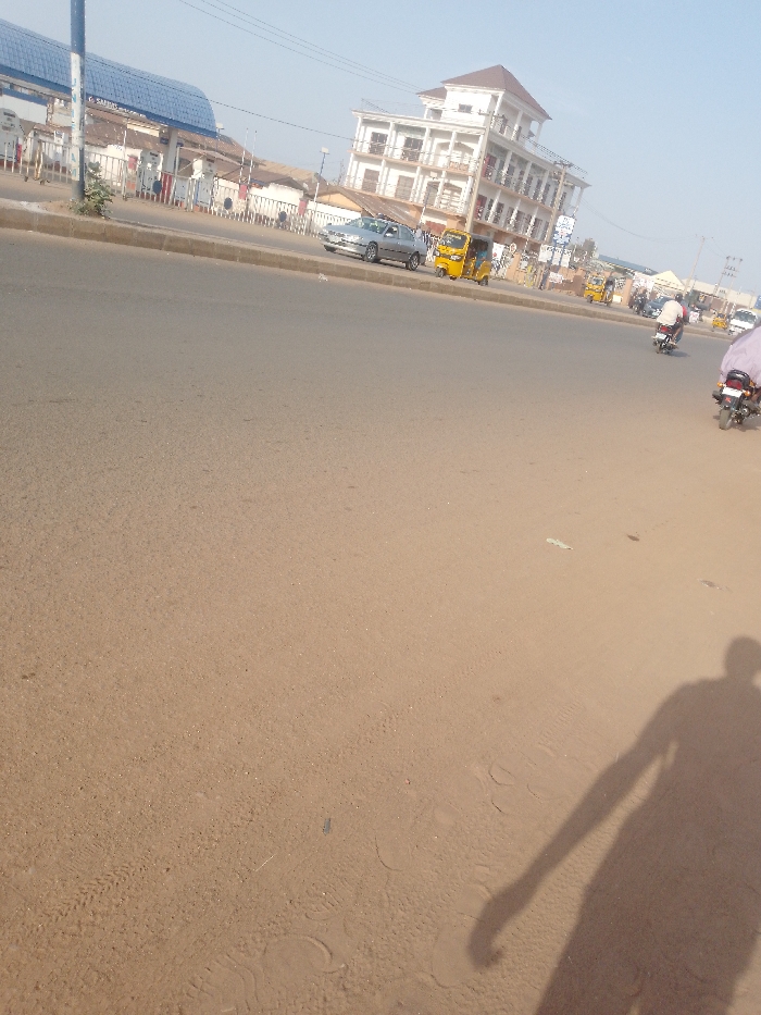 The scene depicts a daylight street view in Sabon Tasha, Nigeria. The foreground consists of a broad expanse of light brown, sandy ground, covering approximately two-thirds of the image from the bottom. A long, dark shadow of a human figure is cast from the lower right corner, extending towards the upper left, onto this sandy surface. Faint footprints and tire tracks are present on the ground.
An asphalt road occupies the middle ground, stretching horizontally across the image. On the far side of this road, a silver sedan is stationary or parked, facing left. Adjacent to it, a yellow motorized tricycle (Keke Napep) is also visible. Further along the road to the right, multiple motorcycles are present, with at least two individuals riding them. One rider, wearing a white garment, is prominently visible on a motorcycle in the right mid-ground, moving away from the viewer.
The background features several structures. On the left, a building with a blue, arched roof is visible, identified as "SAF-RAY," suggesting a filling station with multiple fuel pumps behind a barrier. Towards the center-right, a four-story white building with a brown, pitched roof stands prominently, featuring balconies on each floor. Other smaller buildings, fences, utility poles, and overhead wires are also present along the street line against a clear, light blue sky.