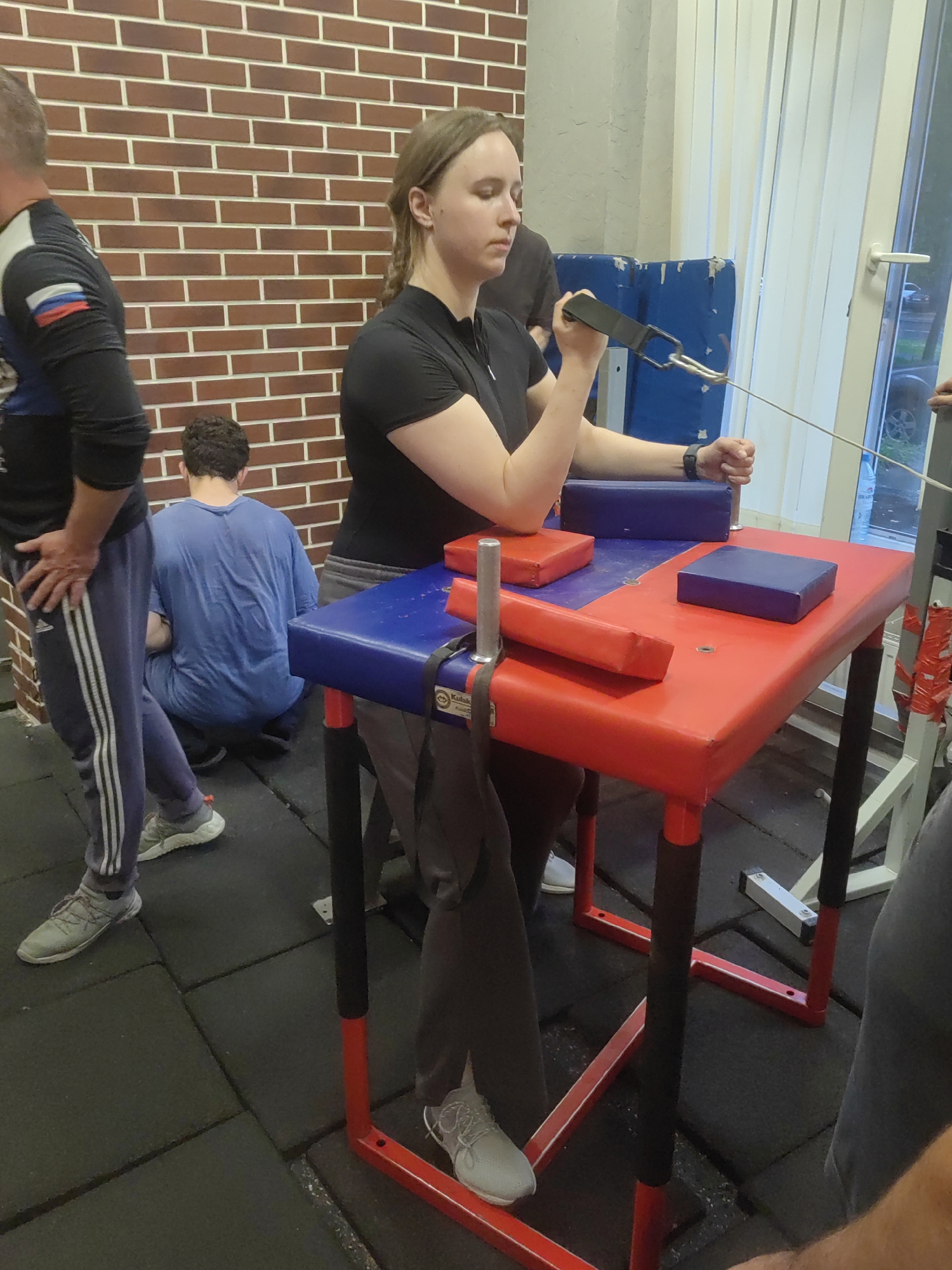 An indoor shot shows a young Caucasian woman with brown hair, wearing a black polo shirt and gray pants, arm wrestling. She is seated at a red and blue arm wrestling table, gripping a black handle attached to a cable that extends out of frame to the right. Her eyes are closed, and her arm is bent at the elbow, suggesting exertion. To her left, a man in a black shirt with a Russian flag patch on the sleeve stands with his left hand on his hip, looking away from the camera. Further left, a person in a blue t-shirt squats against a brick wall. To the right of the arm wrestling table, the lower legs of two individuals are visible. The room appears to be a gym, with black rubber flooring and a brick wall in the background. A large window with vertical blinds is visible to the right. The overall lighting is dim.