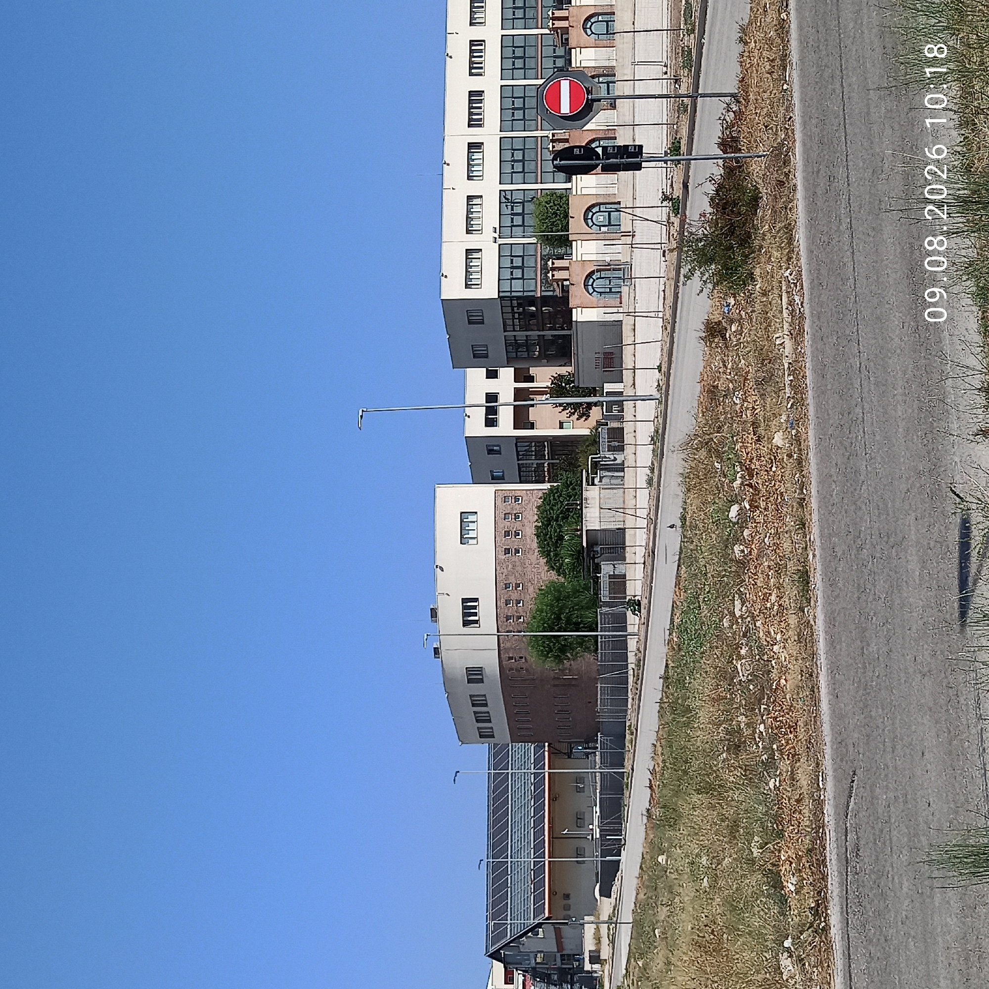 The image shows a modern building complex with a clear blue sky overhead. The buildings appear to be commercial or institutional, with large windows and clean lines. One building has a curved facade and is constructed with a combination of brick and lighter colored materials. Another building, to the left, has a sloped roof covered in solar panels. The area in front of the buildings is somewhat unkempt, with dry grass and some scattered debris. A road runs in the foreground, and a fence separates the road from the property. A "stop" sign is visible near the buildings, mounted on a pole. The timestamp "09.08.2026 10:18" is visible in the lower right corner of the image, suggesting it was taken on August 9, 2026, at 10:18 AM. The architecture and urban setting suggest a location in a developed area, likely a city or industrial park. Given the architecture and climate indicated by the dry vegetation, it is plausible this image depicts a location in Molfetta, Italy, known for its modern infrastructure and sunny Mediterranean climate.