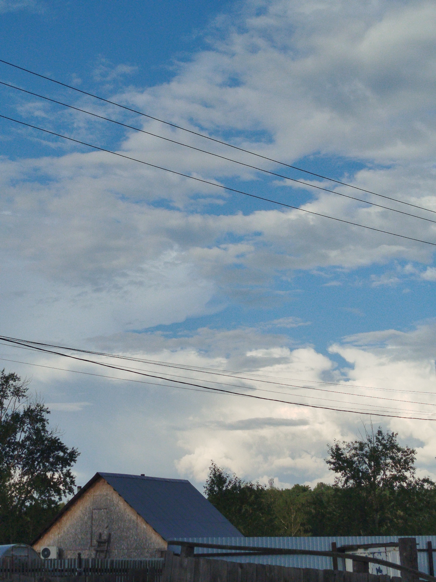The image depicts a rural scene under a cloudy sky with visible power lines in the foreground. A gable-roofed building, possibly a house or shed with a dark metal roof, is situated behind a wooden fence. To the left of the building, an air conditioning unit is attached to the wall. Trees are visible on either side of the building and in the background, forming a dense treeline. The overall impression is one of a calm, somewhat overcast day in a semi-urban or rural environment. This scene is located in Zavodoukovsk, Russia.
