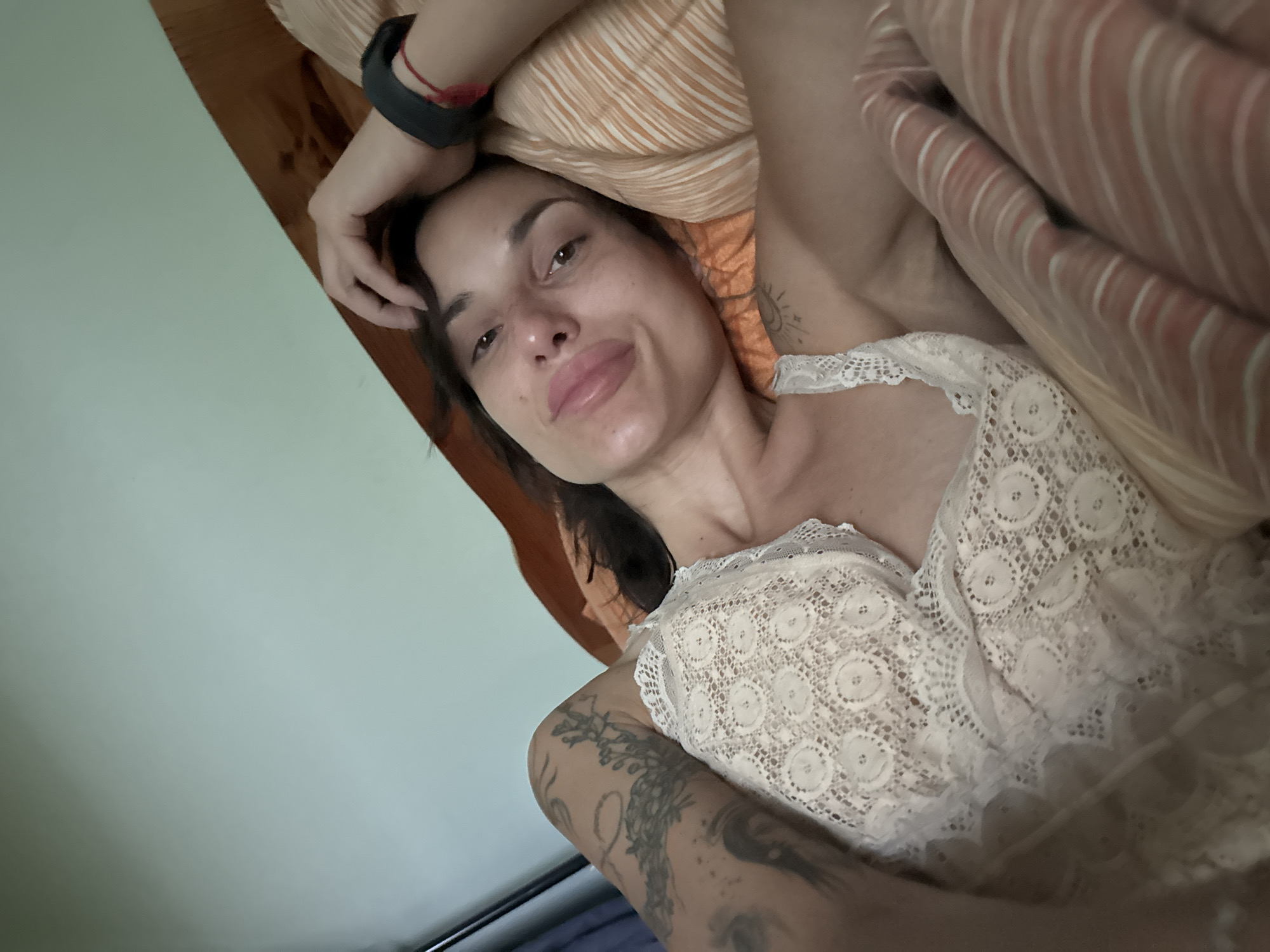 A woman with dark hair, wearing a white lace top, is lying in bed with her head resting on a pillow. She has tattoos on her arm and shoulder. The room appears to be a bedroom, with a wooden headboard and a light-colored wall. The image is a close-up, with the woman's face and upper body filling most of the frame. The lighting is soft and natural.