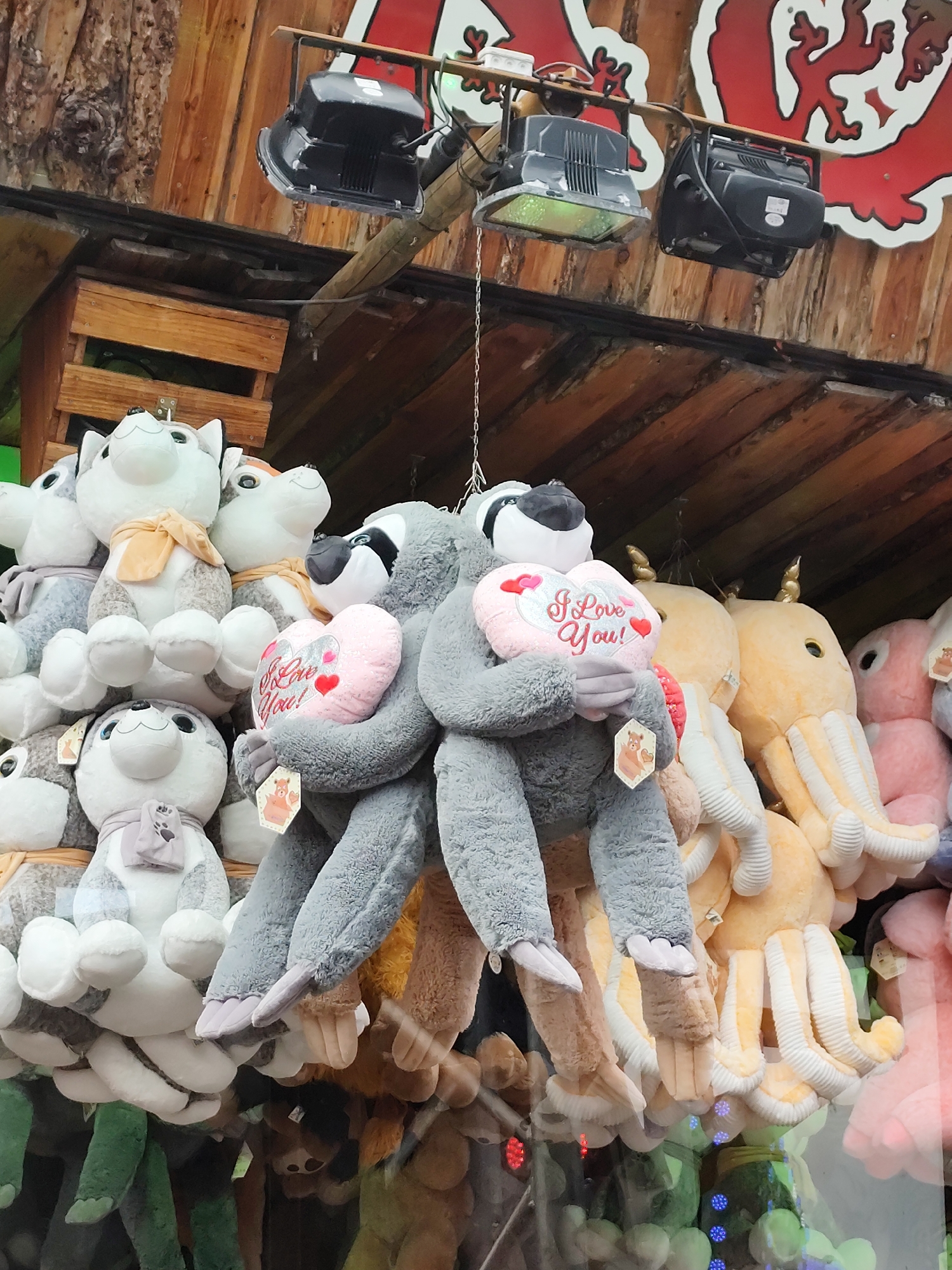 This image shows a display of stuffed animals at an outdoor carnival in Tilburg, Netherlands. The stuffed animals are hanging from a wooden structure and are illuminated by spotlights. The animals include sloths, dogs, and octopuses. The scene appears to be set up for a carnival game, where people would attempt to win the stuffed animals by playing a game. The background shows wooden beams and a red and white banner with a decorative element. The lighting is artificial, suggesting the image was taken at dusk or night.