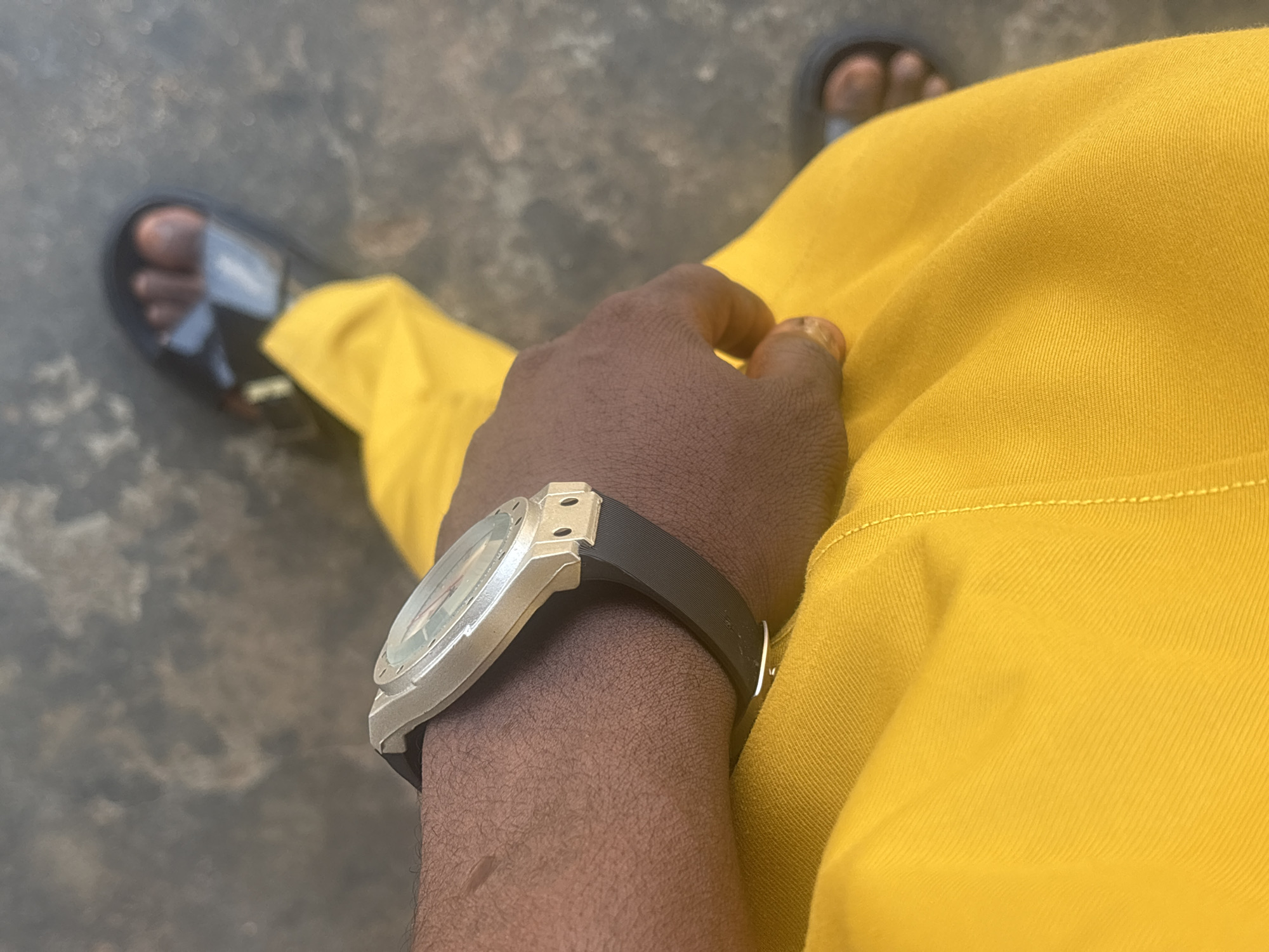 A first-person, high-angle perspective shows a person wearing a silver-colored watch with a black band on their left wrist. The person is wearing a bright yellow garment. The person is also wearing open-toed black sandals. The background is a gray concrete surface. The location is most likely a street in Uyo, Nigeria.