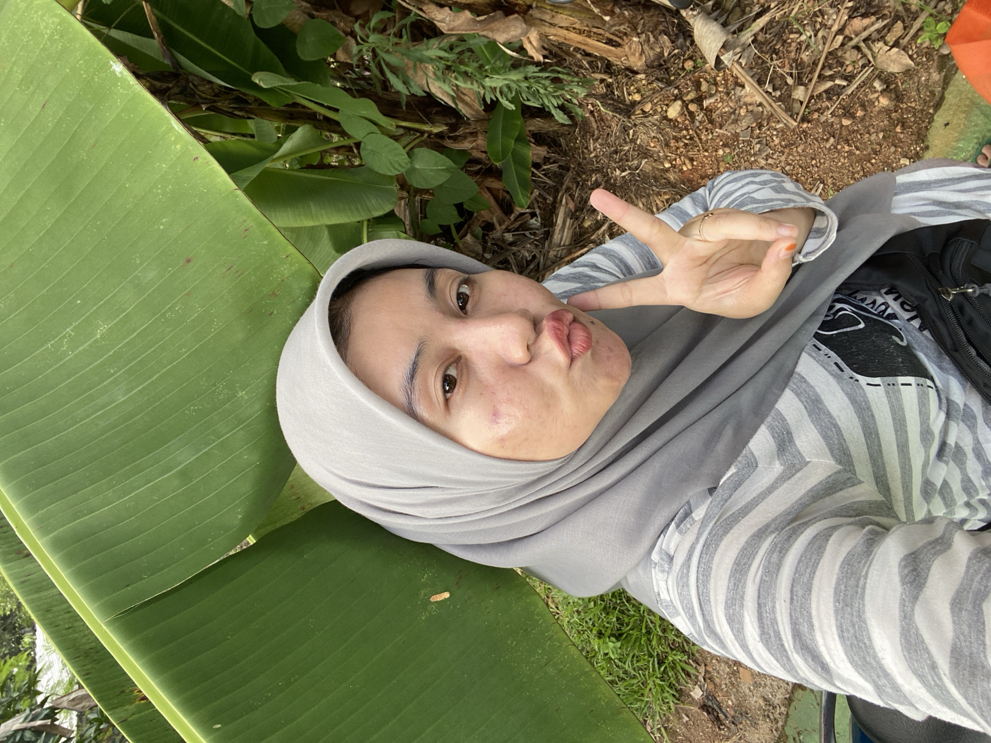 A woman wearing a grey hijab and a striped shirt is taking a selfie outdoors. She is making a peace sign with her right hand and puckering her lips. Behind her are large green banana leaves, some dry leaves, and tree trunks. The ground is covered with dirt and small stones. The scene appears to be in a natural setting, possibly a garden or park. The woman is looking directly at the camera.