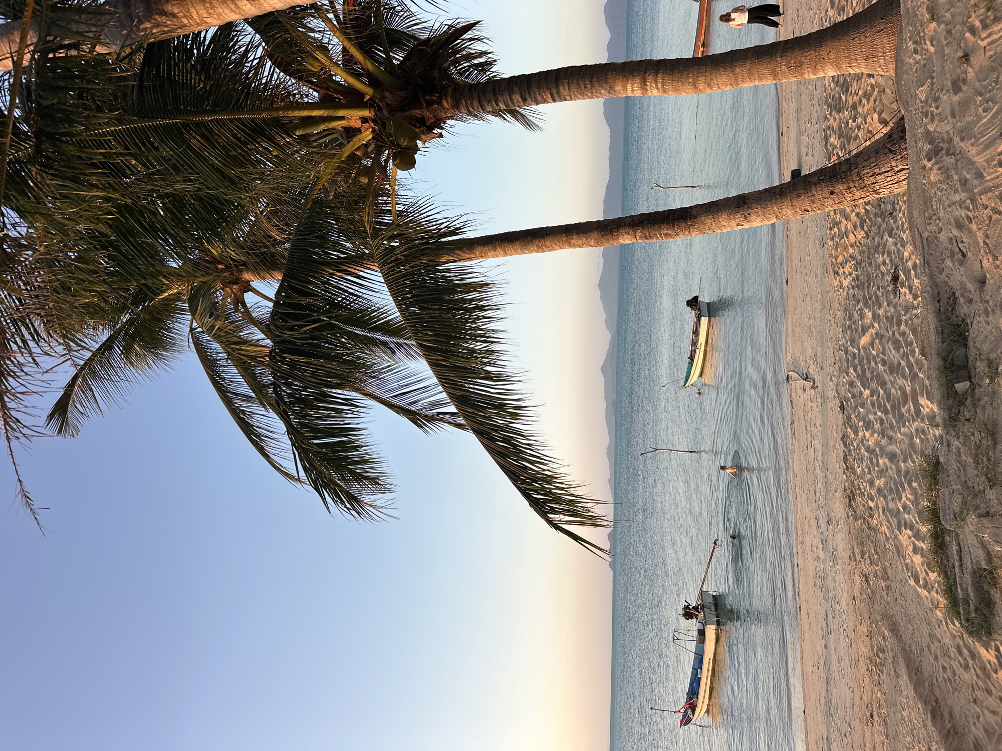 This is a photograph of a beach in the Ko Samui District of Thailand at sunset. Two palm trees are in the foreground, with their trunks partially obscuring the view of the ocean and beach. The sand is light brown and has footprints on it. The ocean is calm and a light blue color, with gentle waves lapping at the shore. In the water, there are two small boats and three people swimming. One boat is white with a red trim and an outboard motor. The other boat is green. In the distance, there are faint outlines of mountains on the horizon. A woman in a white shirt and black pants stands on the beach, facing away from the camera. The sky is clear and a pale blue, with hints of orange and pink from the setting sun.