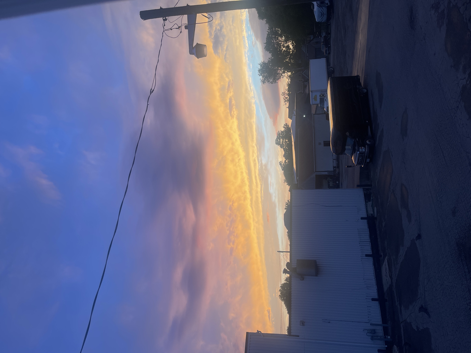 The image shows a wide shot of an industrial area at sunset, with a vibrant sky filled with hues of orange, pink, and purple. In the foreground, a dark-colored van is parked in a paved lot. To the left, a white building with vertical siding is visible. In the background, several other structures, including a large white truck and trees, are silhouetted against the colorful sky. A telephone pole with a light fixture stands on the right side of the frame. The overall scene suggests a commercial or industrial setting during the twilight hours.
