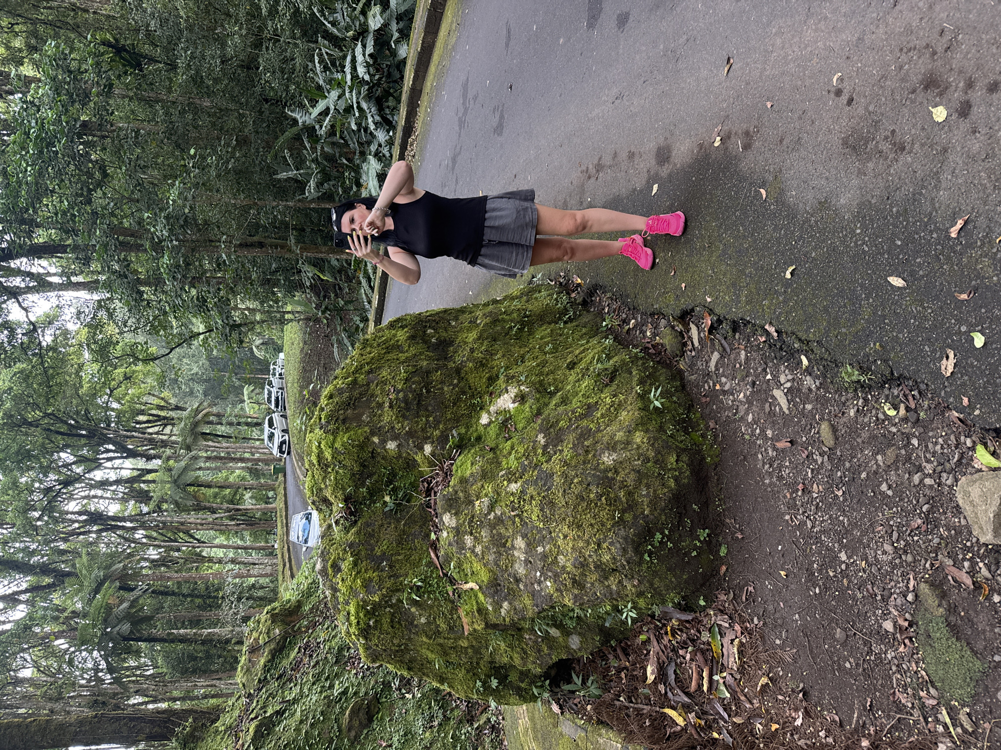 A woman stands on a paved path in a natural setting in Baturiti, Indonesia, using a smartphone to take a selfie. She is wearing a black tank top, grey shorts, and bright pink sneakers. She has dark hair and is wearing sunglasses on top of her head. To her left, a large moss-covered rock sits along the edge of the path. Beyond the rock, a road is lined with tall trees, and several cars are parked along the road. The environment suggests a park or forested area. The sky is overcast.