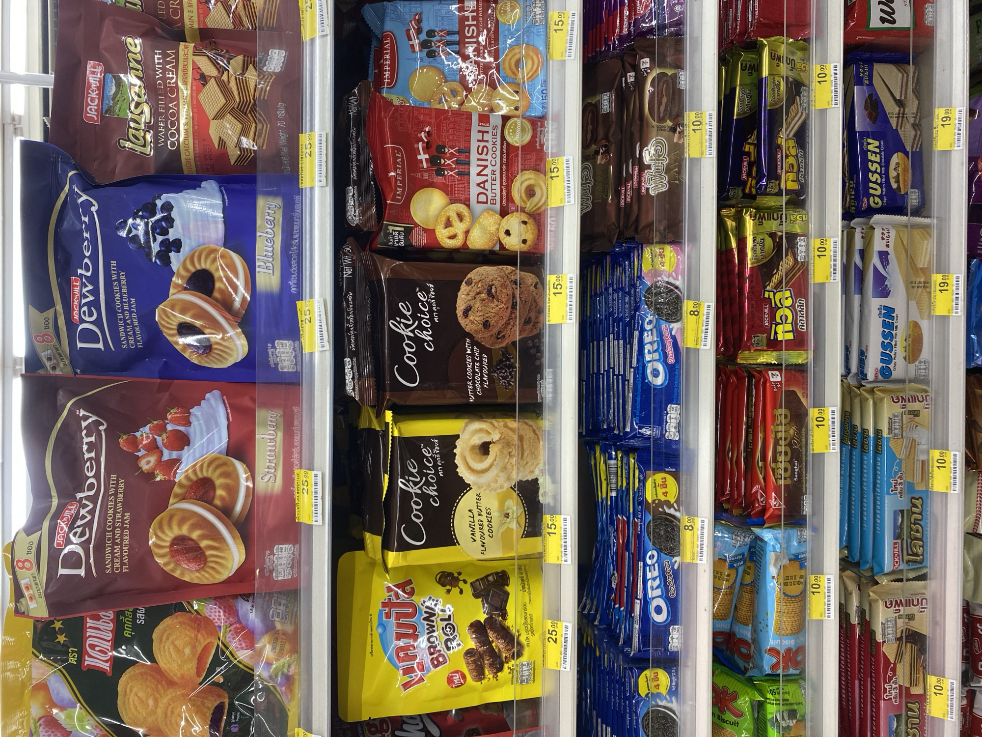 The image displays a well-stocked refrigerated or cooled display case in a store, likely a convenience store or supermarket in Pattaya City, Thailand. The shelves are filled with various packaged snack foods, predominantly biscuits, cookies, and wafers.
Key brands visible include Jack 'n Jill, with products like "Dewberry Sandwich Cookies" in blueberry and strawberry flavors, "Lausanne Wafer Filled with Cocoa Cream," and "Waffu" wafers. Imperial's "Danish Butter Cookies" are prominently featured in different packaging colors, indicating variations like original and chocolate chip flavored. Oreo cookies, both chocolate creme and strawberry creme, are also present in multiple packs. Other brands include "Cookie Choice" (vanilla and chocolate chip butter cookies), "Sanghai Rich" biscuits, "Campus Brownie Roll," and "Gussen Crispy Wafers" with chocolate and milk cream fillings.
Many packages have yellow price tags displaying prices in Thai Baht: 8.00, 10.00, 15.00, 19.00, and 25.00. Product descriptions are primarily in Thai, accompanied by English text for brand names, flavors, and product types. Notable phrases on the packaging include "HIGH CALCIUM & VITAMIN E," "Net Weight," "ขนาดพกพา" (portable size), and "สินค้าขายดีอันดับ 1" (Best Seller No.1). The products are neatly arranged, suggesting an organized retail environment, with some items like Oreo stacked vertically in trays. No people are visible, and the setting appears to be indoors with artificial lighting.