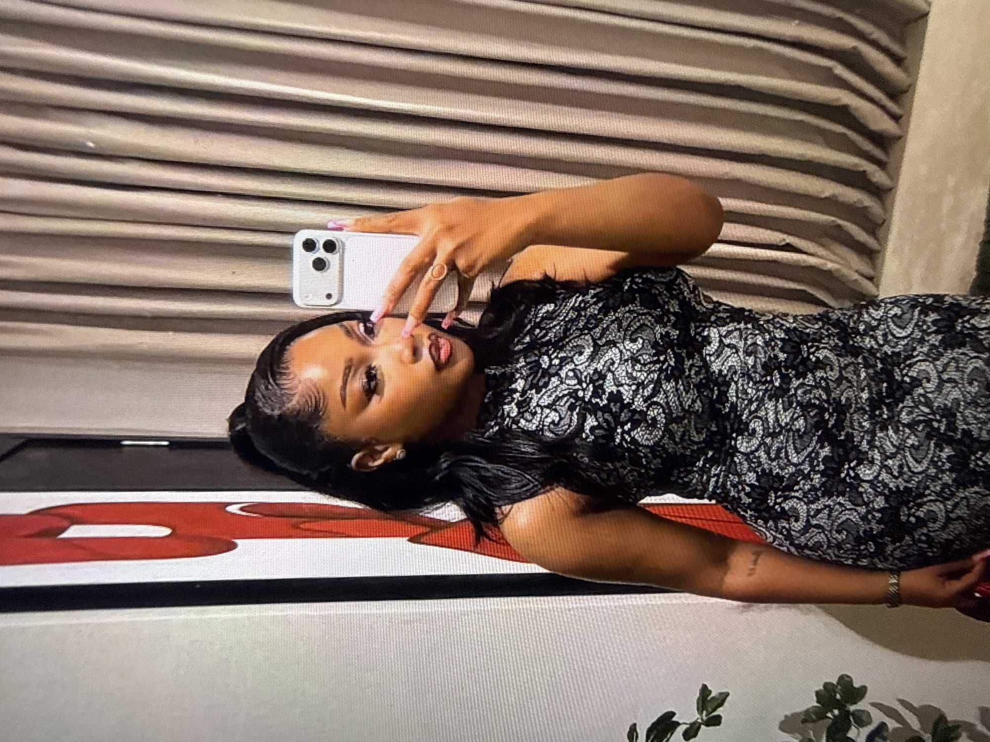 A woman with dark skin, wearing a black and white lace dress, poses for a selfie in a room. She has her hair pulled back in a high ponytail with some strands of hair styled around her face. She is wearing makeup, including eyeshadow and lipstick. Her right hand is holding a white smartphone, and she is wearing a ring on her index finger. Her left arm is bent, with her hand resting on her hip. A portion of a red and white abstract artwork is visible on the left side of the frame, and beige curtains hang in the background on the right. The overall scene appears to be indoors, possibly in a residential or hospitality setting. The woman has a tattoo on her left forearm.