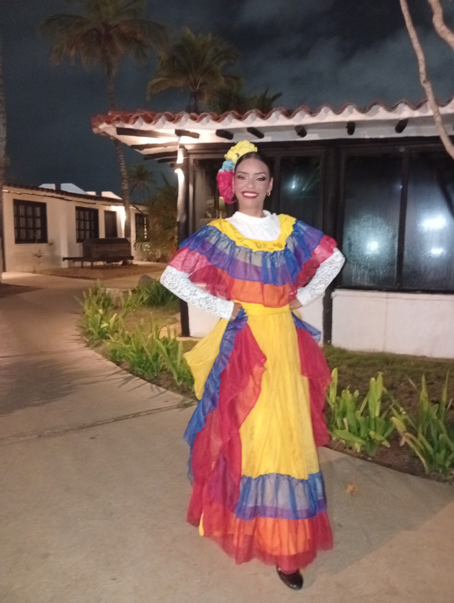 An adult female is standing centrally in a full-body shot on a paved pathway at night in Porlamar, Venezuela. She is smiling directly at the camera, wearing a vibrant traditional costume comprising a white lace-trimmed blouse, a multi-tiered ruffled top in blue, red, and orange, and a long yellow skirt with matching ruffled accents. A floral headpiece with yellow and pink flowers adorns her dark hair. Her hands are positioned on her hips. The background features two low-rise, light-colored buildings with dark-framed windows and reddish-brown tiled roofs. Palm trees are silhouetted against the dark sky. A paved path leads into the background, bordered by green foliage. A dark wooden bench is visible to the left. The scene is illuminated by artificial light sources.