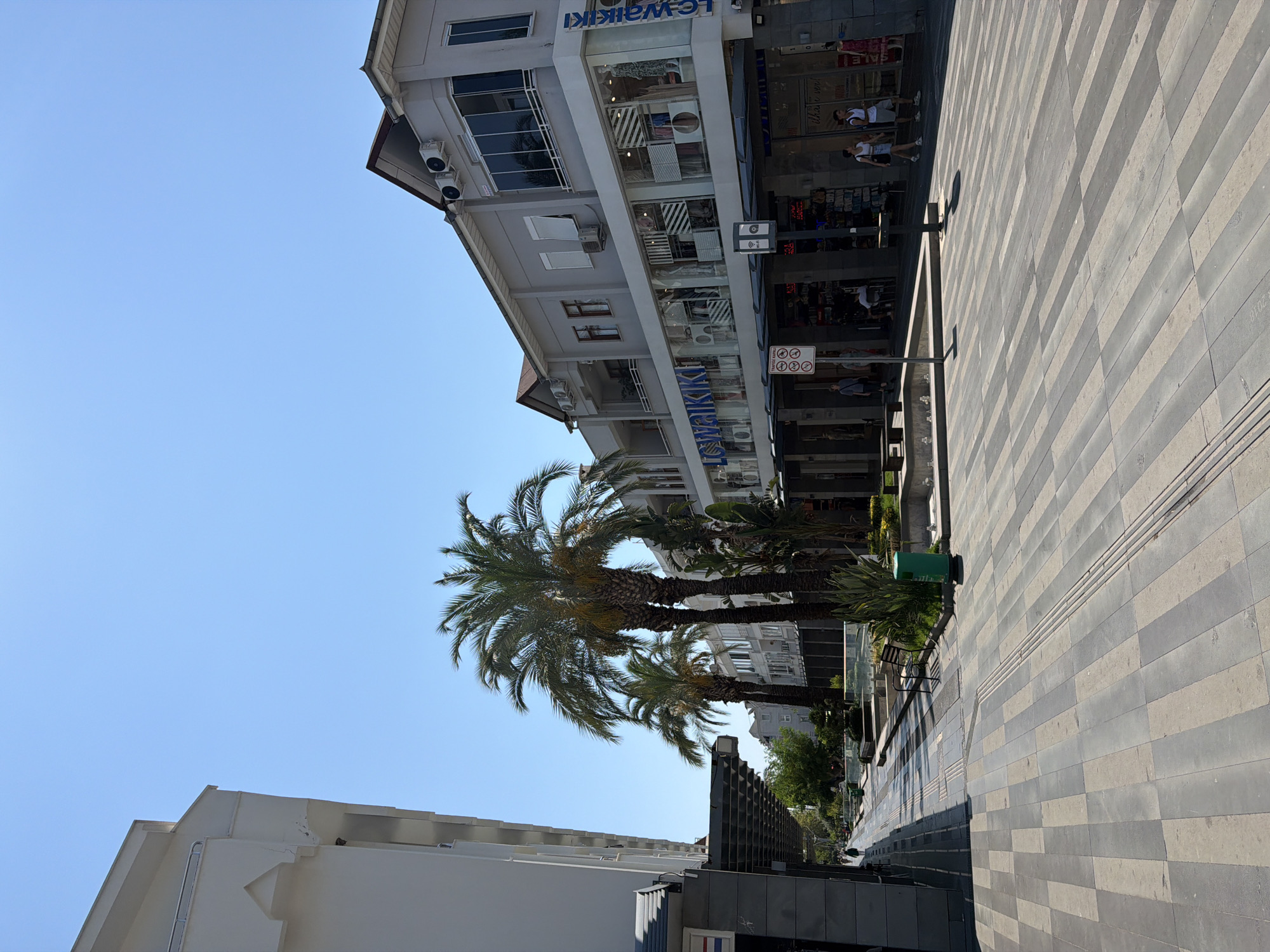 The image depicts a pedestrianized street in Kemer, Turkey, under a clear blue sky. On the right side of the street, a multi-story building houses various shops, prominently featuring a large LC Waikiki store. The building's facade is light gray, with large glass windows displaying merchandise and mannequins. Air conditioning units are visible on the upper levels. Palm trees line the street, casting shadows on the patterned pavement. On the left side of the street, another building, primarily white, is visible, with awnings extending over a walkway. Several individuals are walking along the street, some browsing shop windows. Green planters with vegetation and benches are interspersed along the walkway, suggesting a public area designed for leisure. The overall scene conveys a sunny day in a commercial district.