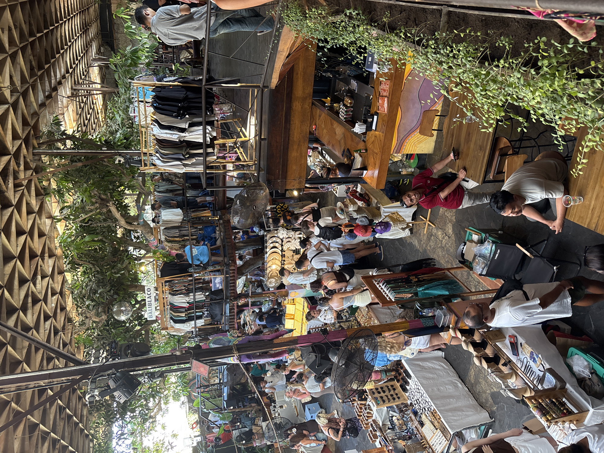 The image depicts a bustling indoor market, likely within Kuta Selatan, Indonesia, with a rustic, open-air design featuring a wooden latticed ceiling and exposed beams. The market is multi-leveled, with walkways and stalls arranged on different tiers. On the upper level, racks of clothing are displayed, and a disco ball hangs from the ceiling amidst lush greenery. People are seen browsing at clothing stalls, and some are seated on stools.
The middle level features more clothing racks, and walkways where people are walking and shopping. On the lower level, various market stalls are set up. One stall prominently displays jewelry and accessories on tables covered with white cloths. Another stall shows handmade crafts and other goods. People are actively engaged in shopping, examining items, and interacting with vendors.
A bar or cafe area is visible on one side, with wooden countertops and seating. Several individuals are seated at tables in this area, one is standing at the bar and another is looking at merchandise. The overall atmosphere is lively and crowded, characteristic of a popular marketplace. The presence of tropical plants and the architectural style suggest a location in a warm climate, consistent with Kuta Selatan, Bali, Indonesia.