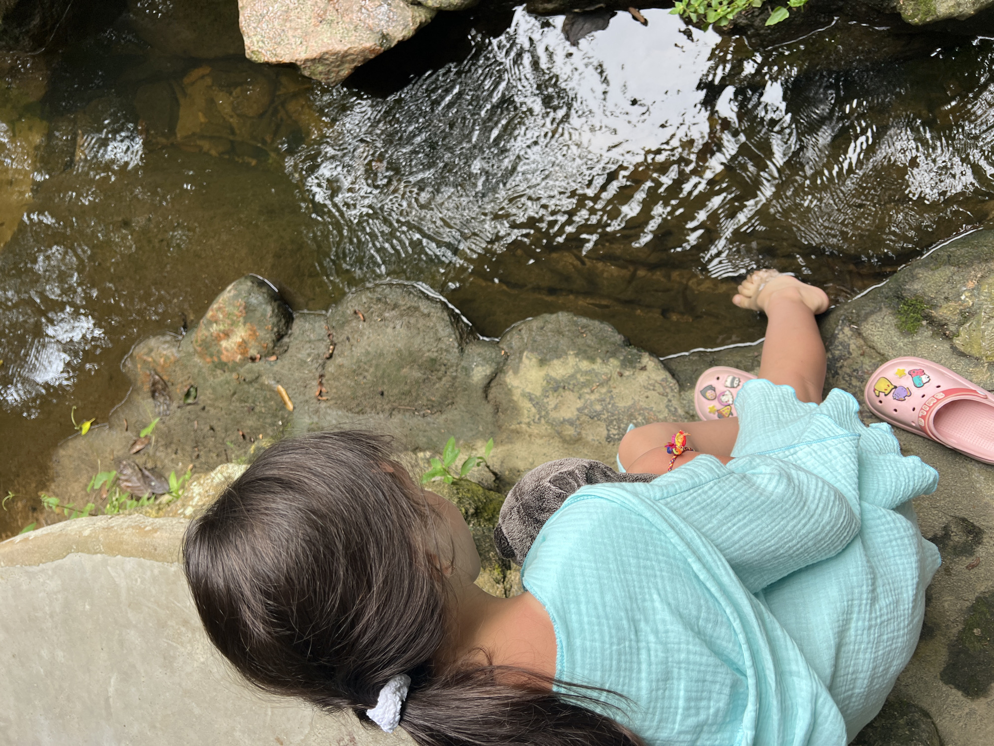 This media file shows a young girl from the shoulders up, seated on a concrete ledge next to a shallow, clear stream in Ko Samui, Thailand. The water flows over rocks, with some greenery visible on the banks. The girl has dark, long hair tied back in a ponytail. She is wearing a light blue, crinkled fabric garment, possibly a dress or a robe. Her right foot is partially submerged in the stream, and her left foot is visible in a pink slip-on sandal decorated with cartoon characters. Another pink sandal is beside her left foot. The overall scene suggests a warm, possibly humid day, common in Thailand. The girl's posture and focus on the stream suggest a moment of quiet observation or play. The lighting indicates it is daytime, with natural light filtering through. No text is visible in the image.