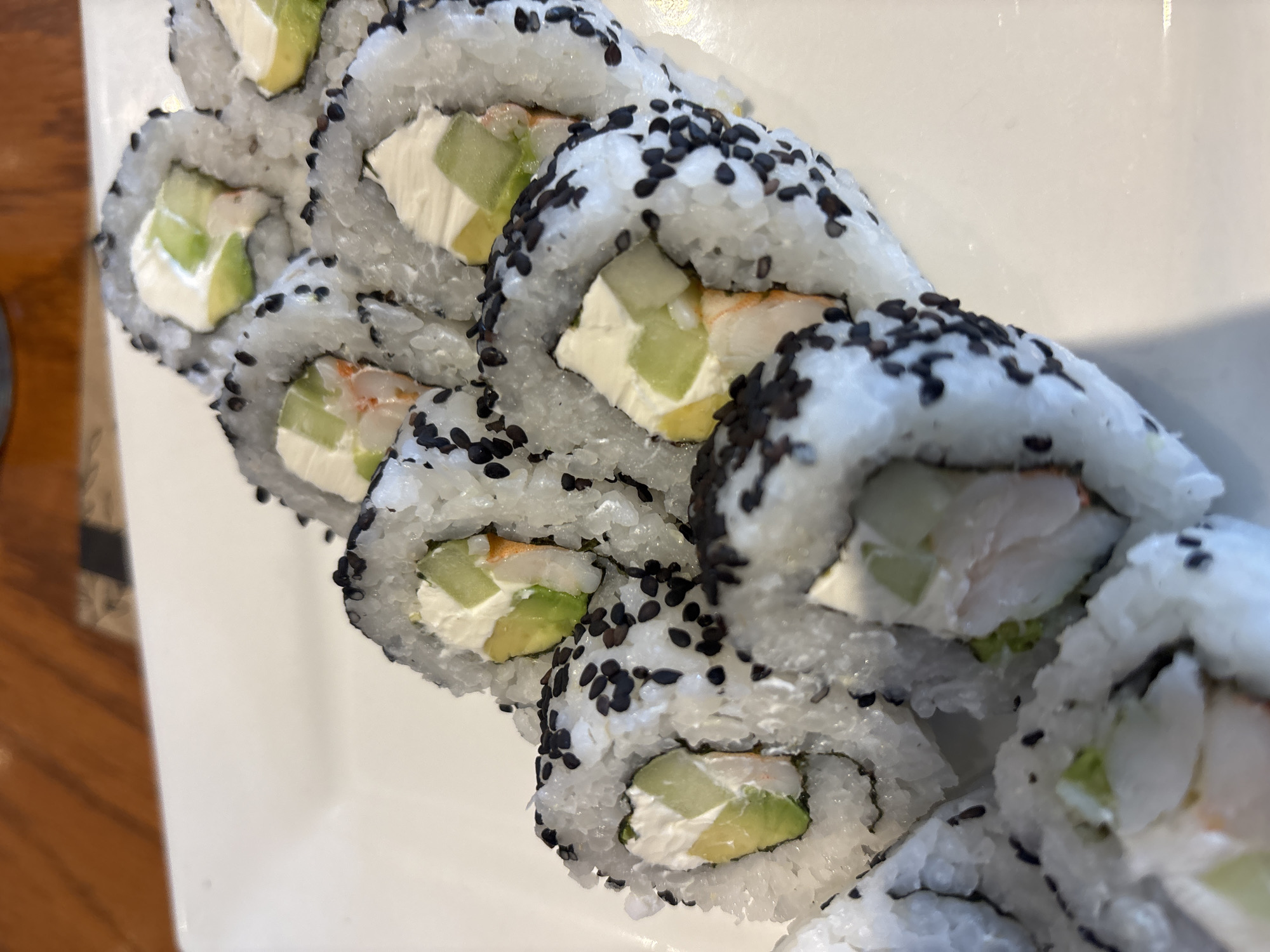 A close-up shot captures a row of neatly sliced sushi rolls arranged on a clean, white rectangular plate. The sushi rolls, presented as uramaki (rice on the outside), are generously coated with black sesame seeds, giving them a speckled appearance against the white sushi rice.
Each roll's cross-section reveals its vibrant filling: creamy white cream cheese, bright green slices of avocado, light green rectangular pieces of cucumber, and faint hints of pinkish-white meat, likely imitation crab. The rice appears slightly moist and well-formed around the fillings.
The rolls are aligned diagonally across the plate, creating a sense of depth. Part of a warm-toned wooden surface is visible in the background on the left, suggesting the sushi is placed on a table. The lighting is bright and even, highlighting the textures and colors of the food. There are no people visible, nor any discernible text. The image focuses entirely on the prepared sushi, likely ready to be served in Puebla, Mexico.