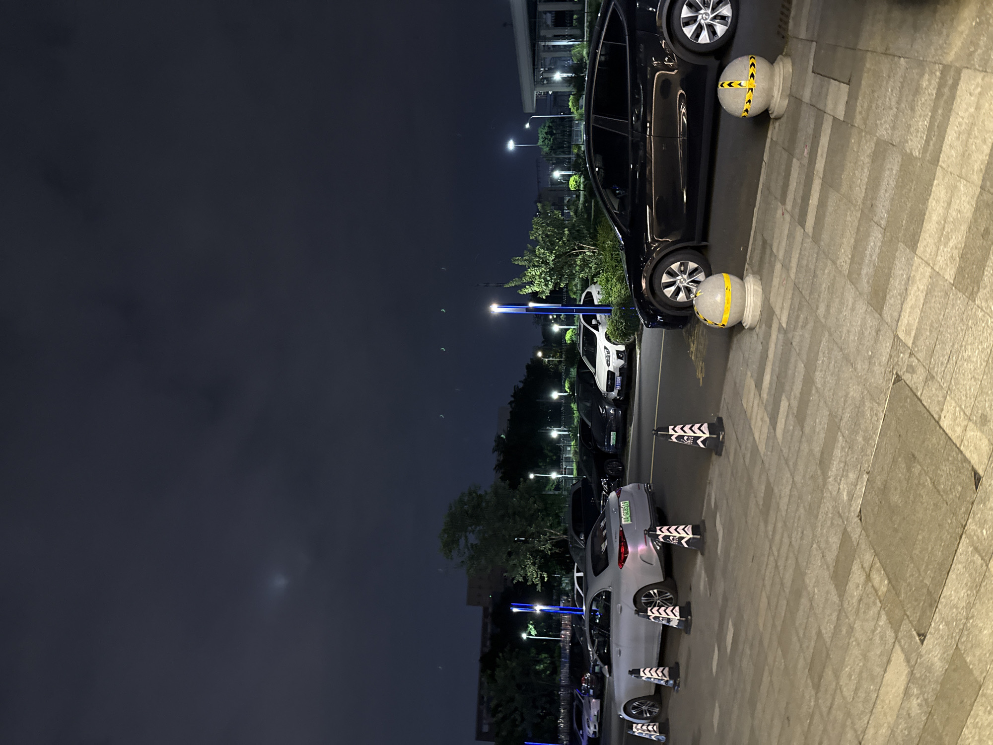 An exterior, ground-level, eye-level, nighttime shot depicts a parking lot scene in Huangpu District, China. The sky is dark with some faint clouds and a visible bright spot, possibly a light reflection or atmospheric phenomenon. Several cars are parked in rows, including a silver sedan in the foreground with a license plate reading "AA GCG519", and a black SUV-style vehicle in the middle ground. Traffic cones are placed along the parking lot's edge, some conical and black and white striped, and two spherical bollards with yellow and black striped tape are present on the paved walkway. The background shows trees, buildings, and streetlights, suggesting an urban or suburban setting. The ground is paved with large, light-colored tiles.