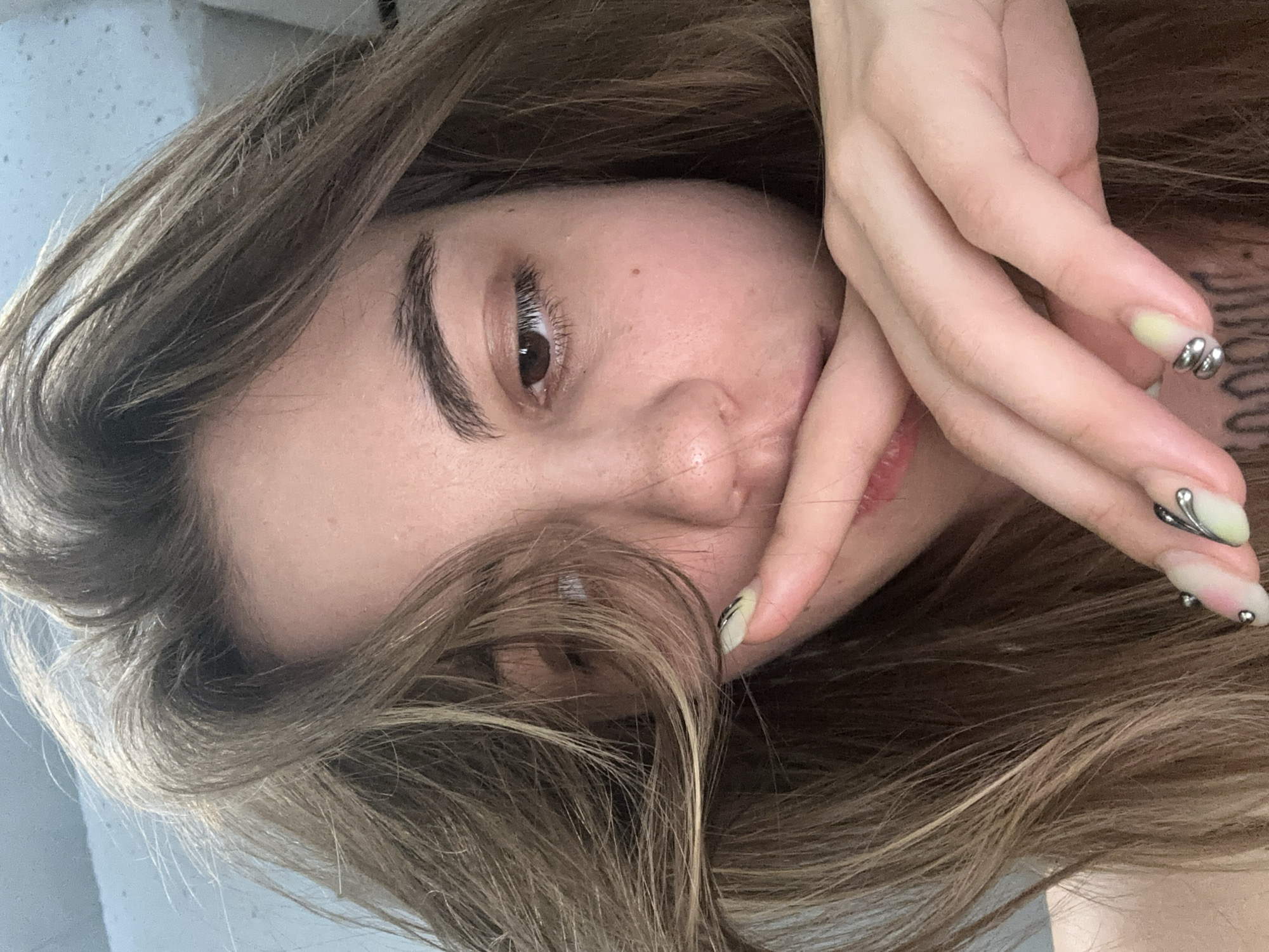 A close-up, low-angle selfie shows a young woman with long, light brown hair covering part of her face. Her hand is raised to her mouth, obscuring her lips and chin. Her right eye is visible, looking towards the left side of the frame. She has fair skin with some freckles and rosy cheeks. Her fingernails are decorated with a pale green and pink gradient and silver embellishments. The background is blurred, appearing to be an indoor setting with a light blue wall. There is no indication of the city of Moscow, Russia, or any specific location within it.